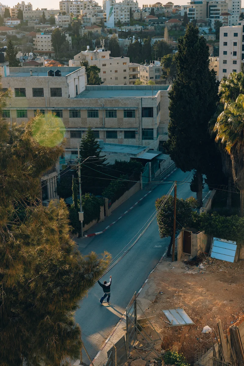 Landing by Maen Hammad, photographed by Maen Hammad — A skateboarding figure navigates a blue-painted street in a Palestinian neighborhood, captured from an elevated vantage point.