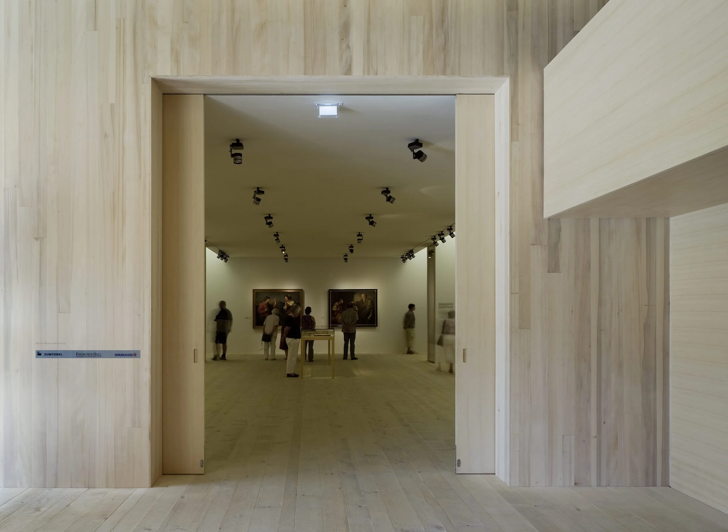 The Angelika Kauffmann Museum by Dietrich Untertrifaller, photographed by Bruno Klomfar — The museum interior reveals a post-and-beam timber structure supporting a corrugated metal roof, with exposed wooden framing and blue steel support columns.