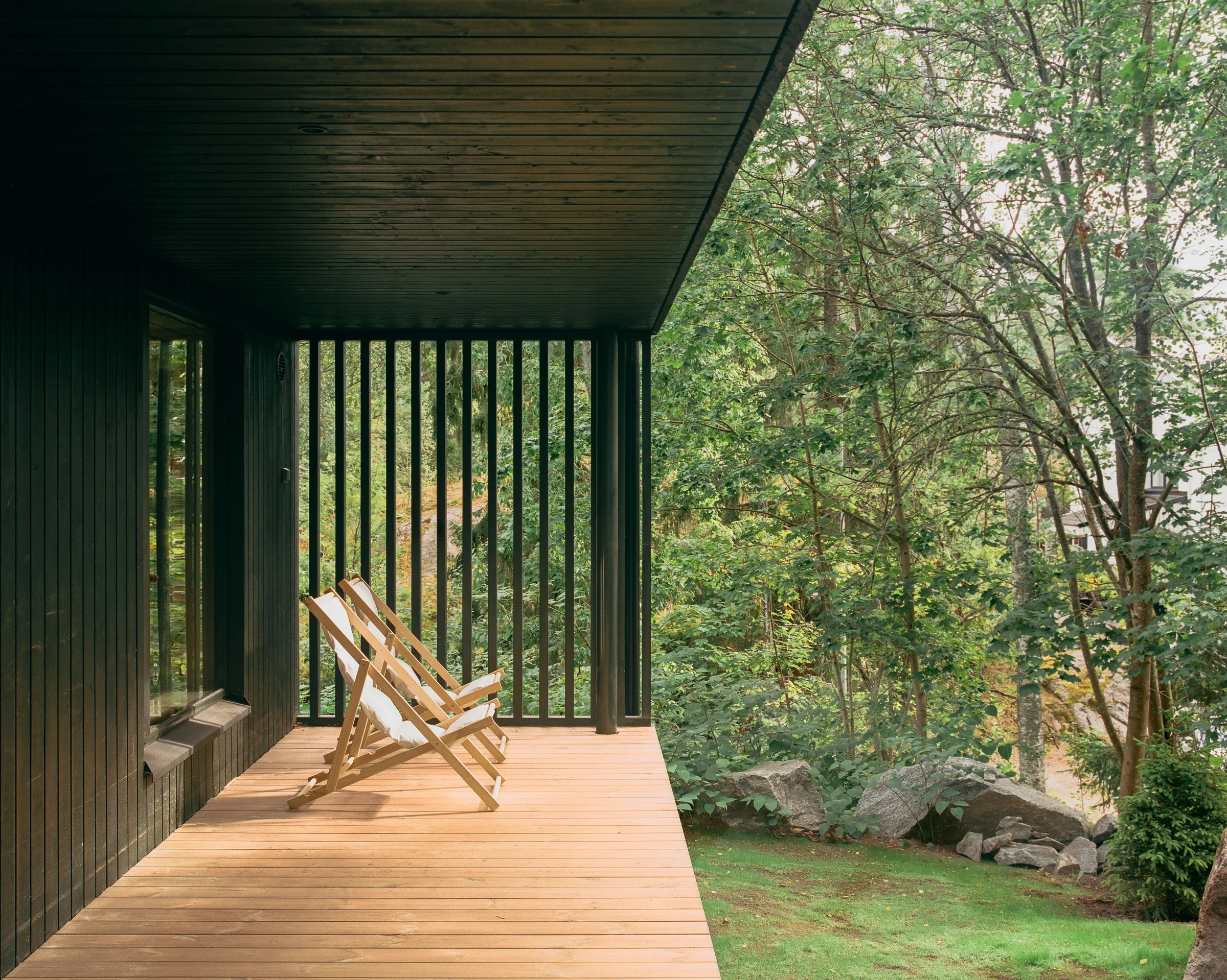 Villa Margo by Talli Ark, photographed by Kalle Kouhia — A hallway in light pine paneling frames two thresholds: a horizontal window opening onto the sloped landscape beyond, and a glazed doorway leading to a covered deck with metal railings.