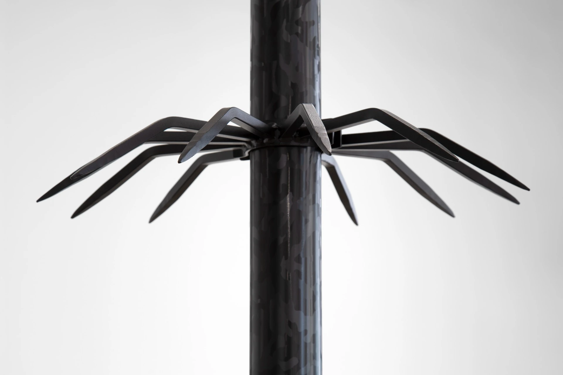 Would you wear my eyes? by Nicolas Lebeau — Detail of Totem a: a camouflage-patterned steel column with a ring of eight welded anti-climb spikes, pointed and splayed outward like a spider at rest.
