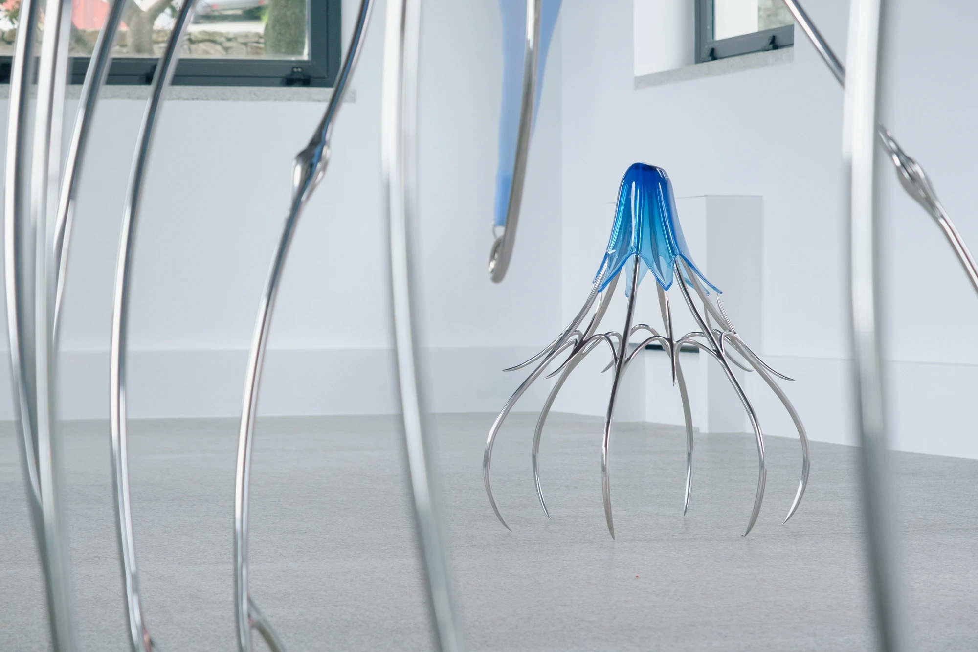 Blue Blooded — Sangue blu by Hannah Levy, photographed by Andrea Mignogna — a cobalt glass bell sits on a ring of hooked steel legs, shot through the forest of another sculpture's legs in the foreground.
