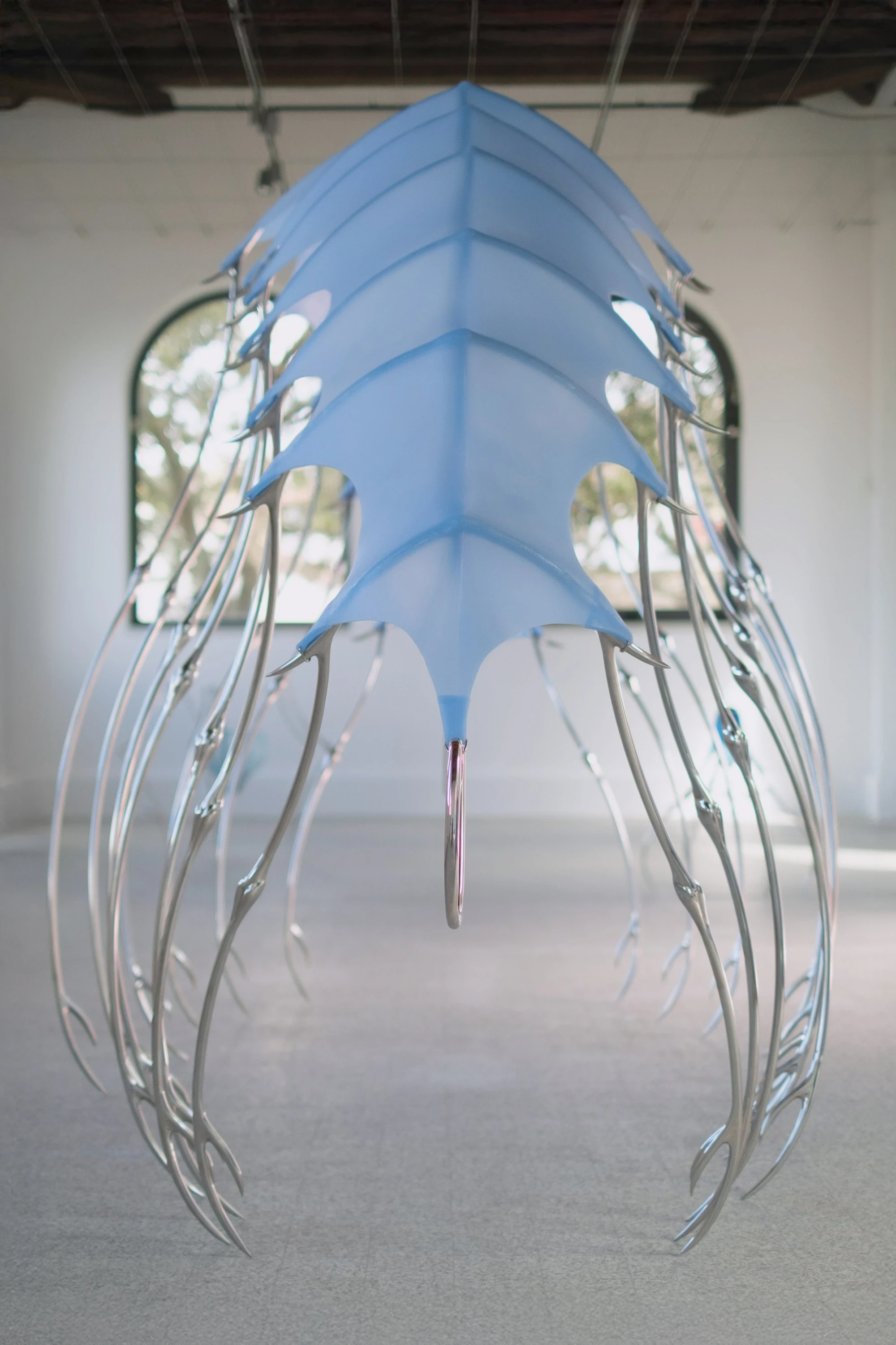 Blue Blooded — Sangue blu by Hannah Levy, photographed by Andrea Mignogna — the central silicone-and-steel canopy hangs head-on in the nave, ribbed like a fossilised carapace with long talon-tipped legs reaching toward the terrazzo floor.