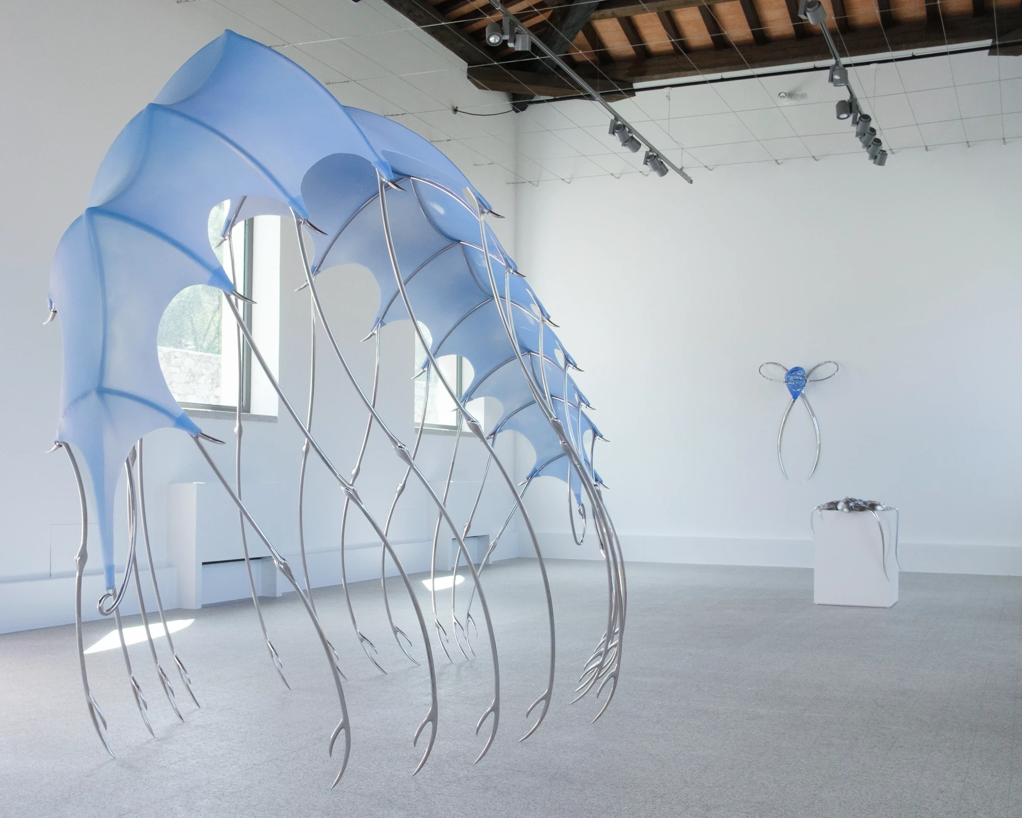 Blue Blooded — Sangue blu by Hannah Levy, photographed by Andrea Mignogna — a three-quarter view of the silicone canopy with a smaller wall sconce holding blue glass visible behind it, the gabled wood ceiling of the washhouse overhead.