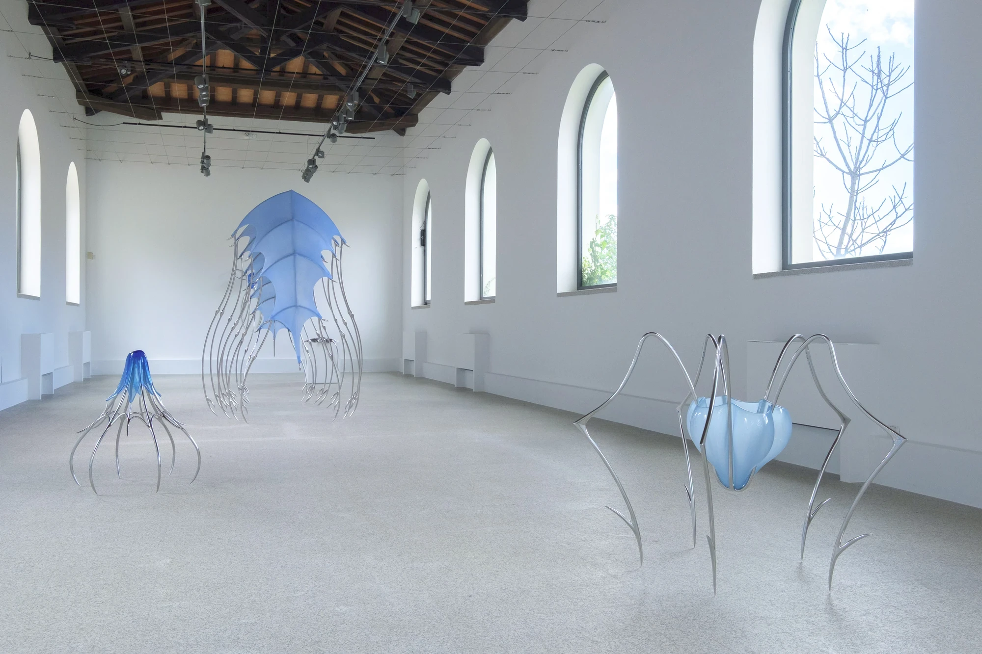 Blue Blooded — Sangue blu by Hannah Levy, photographed by Andrea Mignogna — long installation view down the Museo Nivola nave, five sculptures in steel, silicone and blown blue glass distributed through the barrel of arched windows.