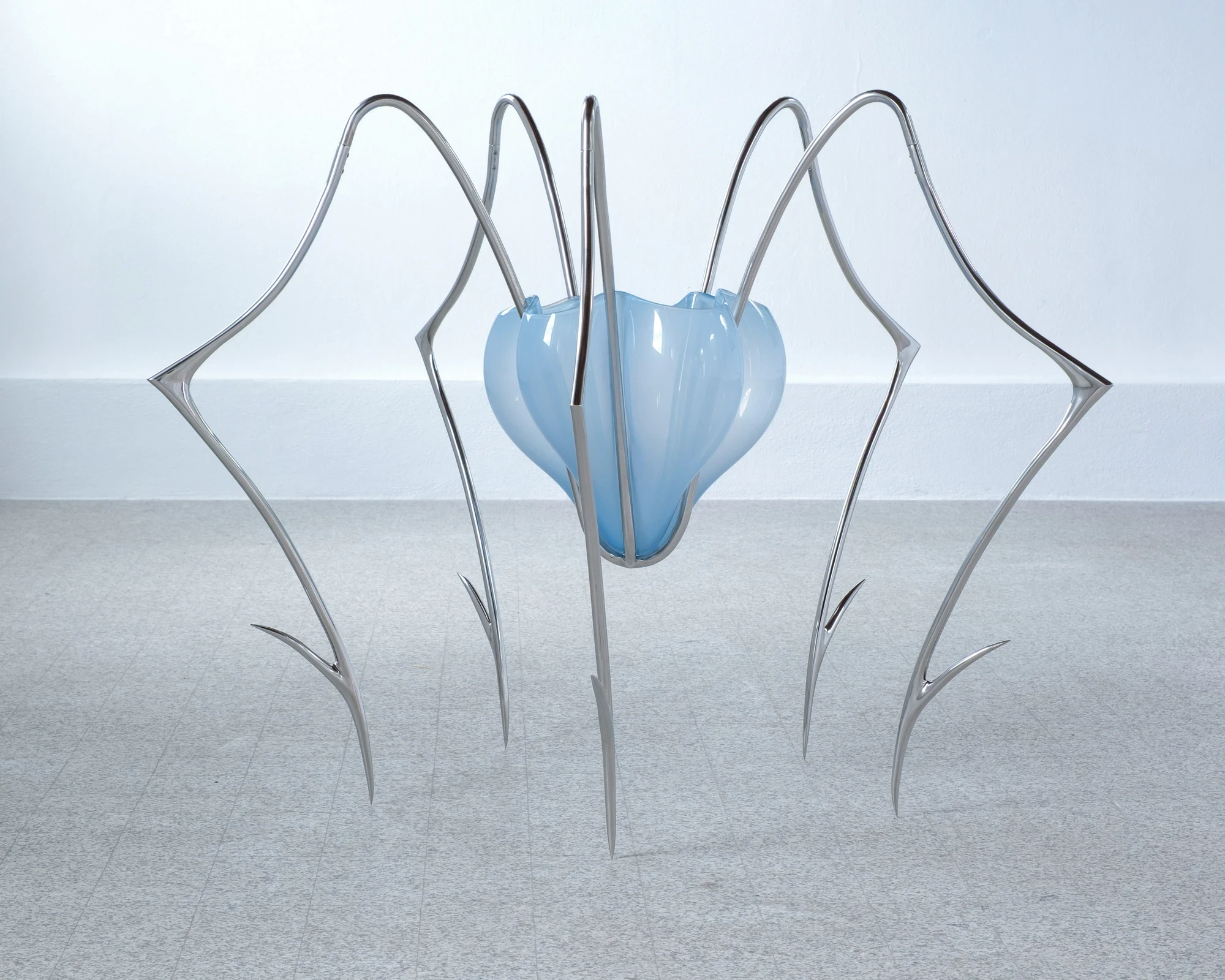 Blue Blooded — Sangue blu by Hannah Levy, photographed by Andrea Mignogna — front-on study of a blown-glass form nested inside a cage of ribbed steel legs whose claw tips splay outward onto the polished floor.