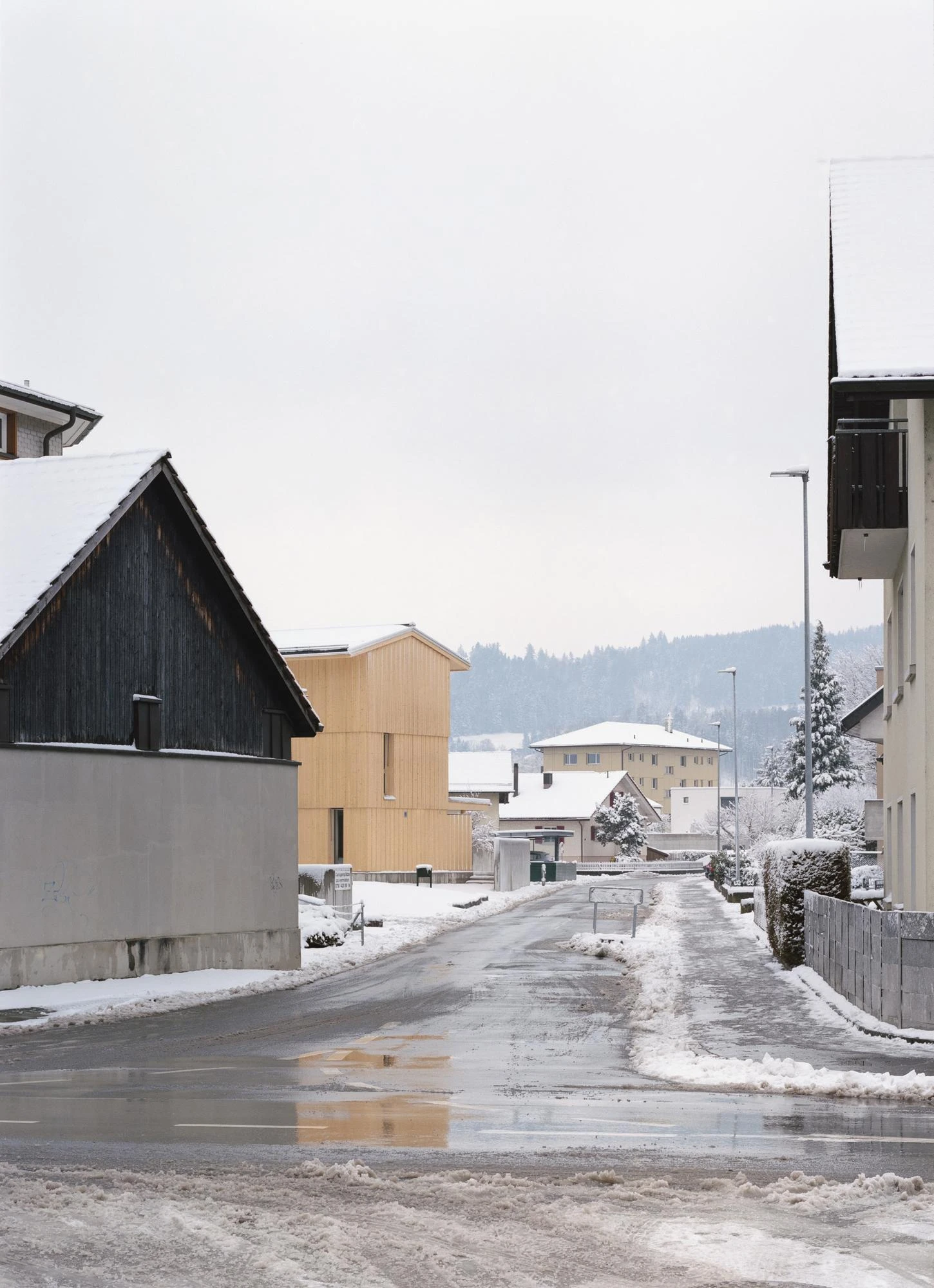 Tower House by kit, photographed by Till Forrer — Front elevation from the street in winter; the three-storey spruce volume against snow-covered shrubs, a small timber outbuilding to the left.