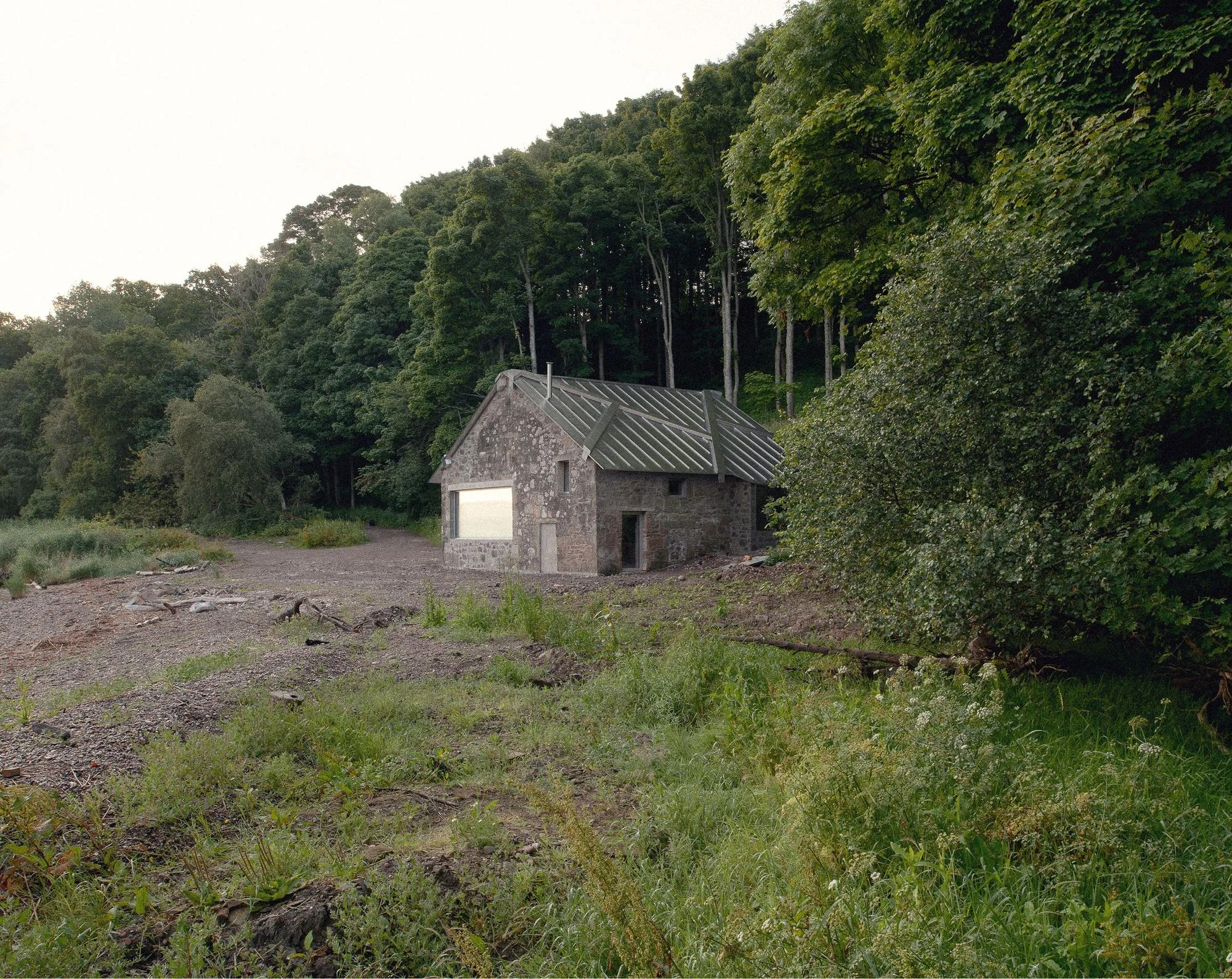 Boathouse by Barboza Blanco Office, photographed by O.C. Photography, Rohan Strathie, Barboza Blanco — Wide view of the completed boathouse from the river bank: stone walls with a corrugated metal roof, glowing interior light in one window.
