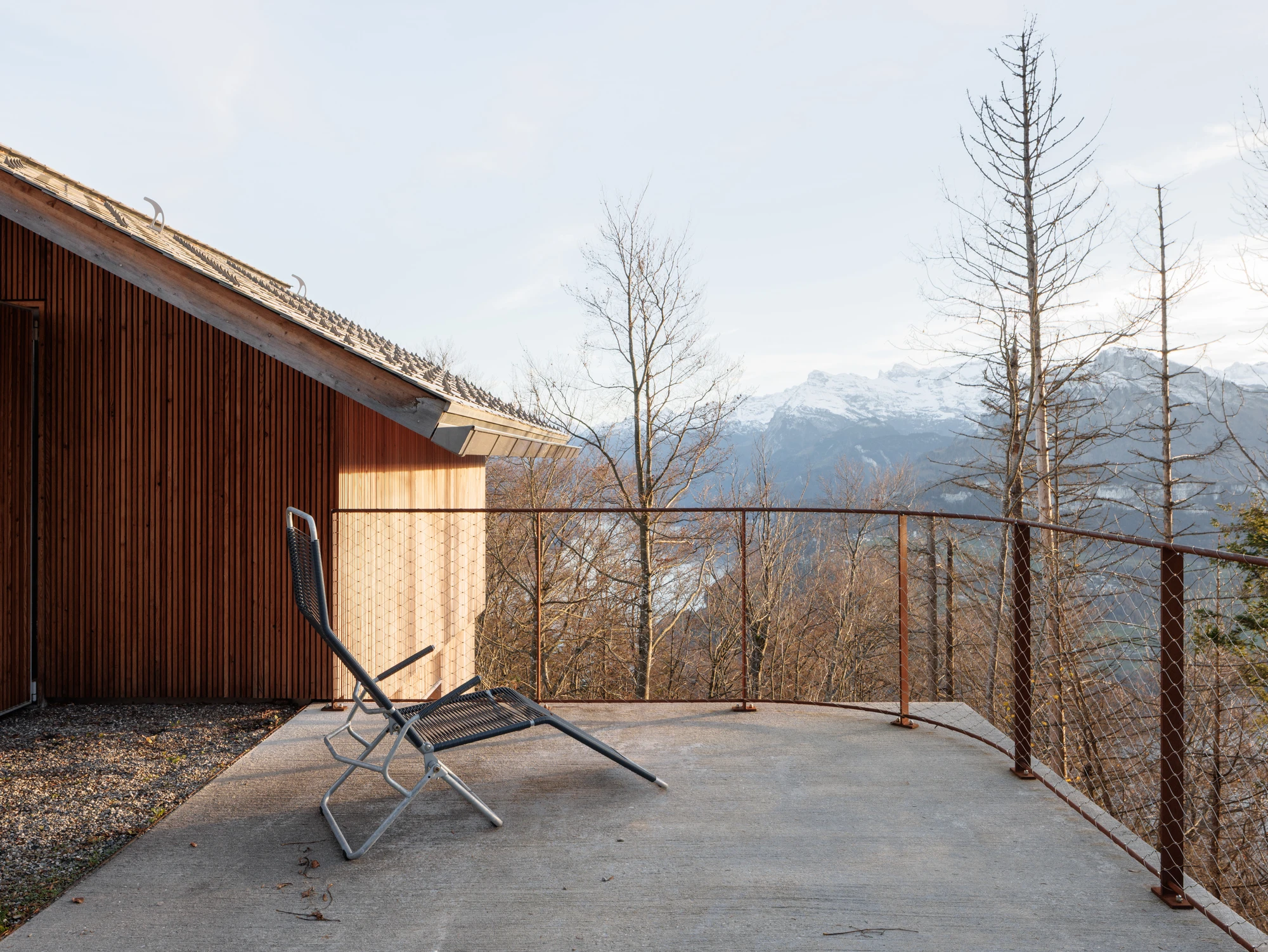 Swiss Alpine Cabin Reconstruction by Guča arch., photographed by Matej Hakár — Exterior terrace view: the raw concrete fireplace with two deck chairs, the Corten rail fence at the edge, Lake Uri and the Alps in the distance.