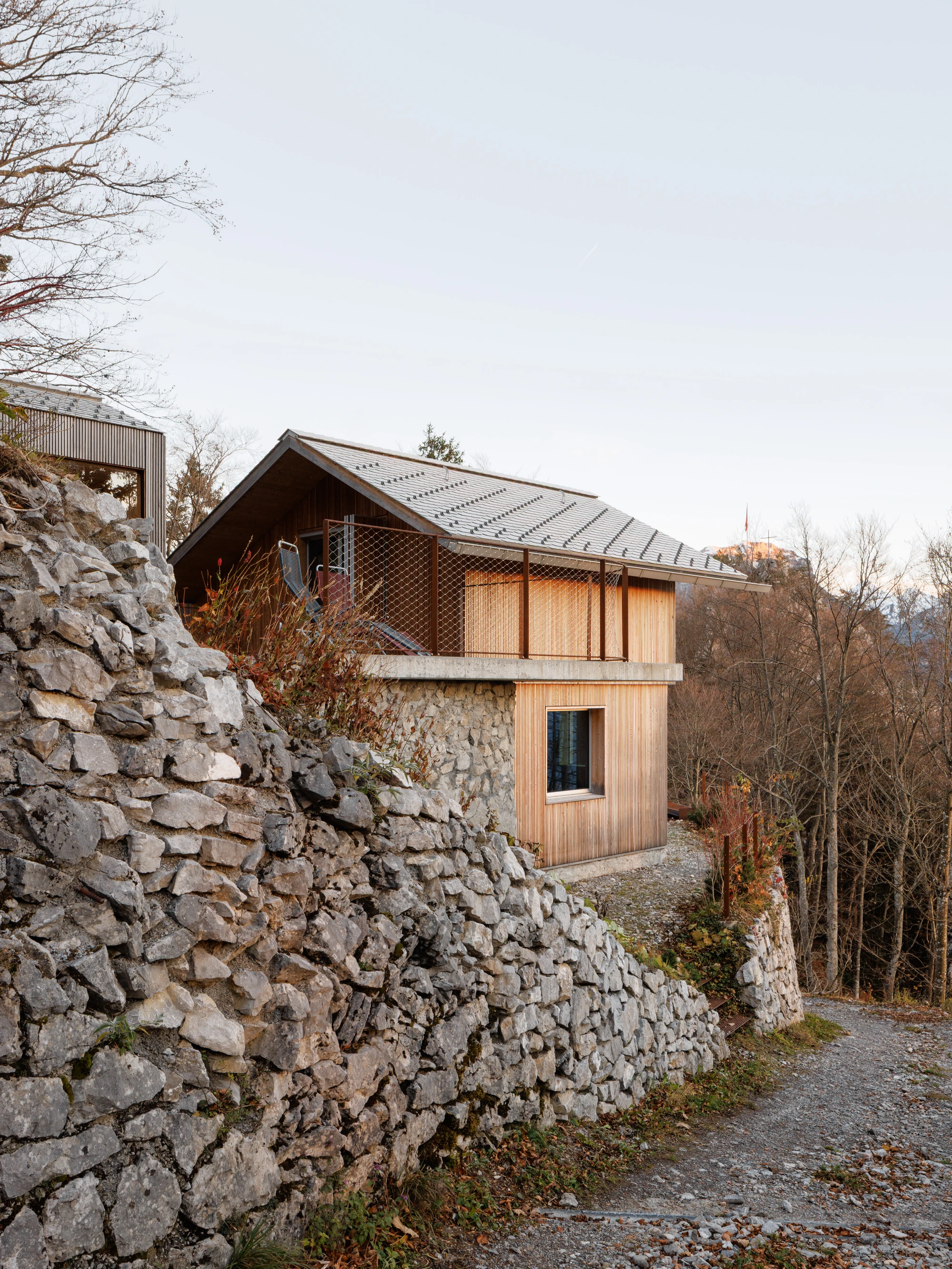 Swiss Alpine Cabin Reconstruction by Guča arch., photographed by Matej Hakár — Interior view from living area toward the large glass facade: rock face close outside, pale CLT interior behind, a walnut table and chair in the foreground.