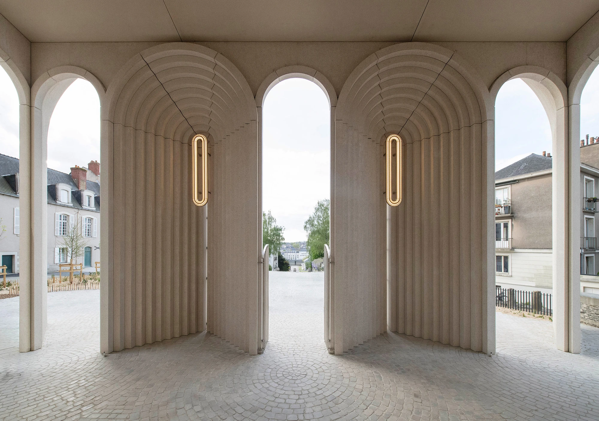 Angers Cathedral Entrance by Kengo Kuma and Associates — Interior view through the gallery piers: slender neon verticals mark the concrete columns while cobblestone and the city beyond are visible through open arched passageways.