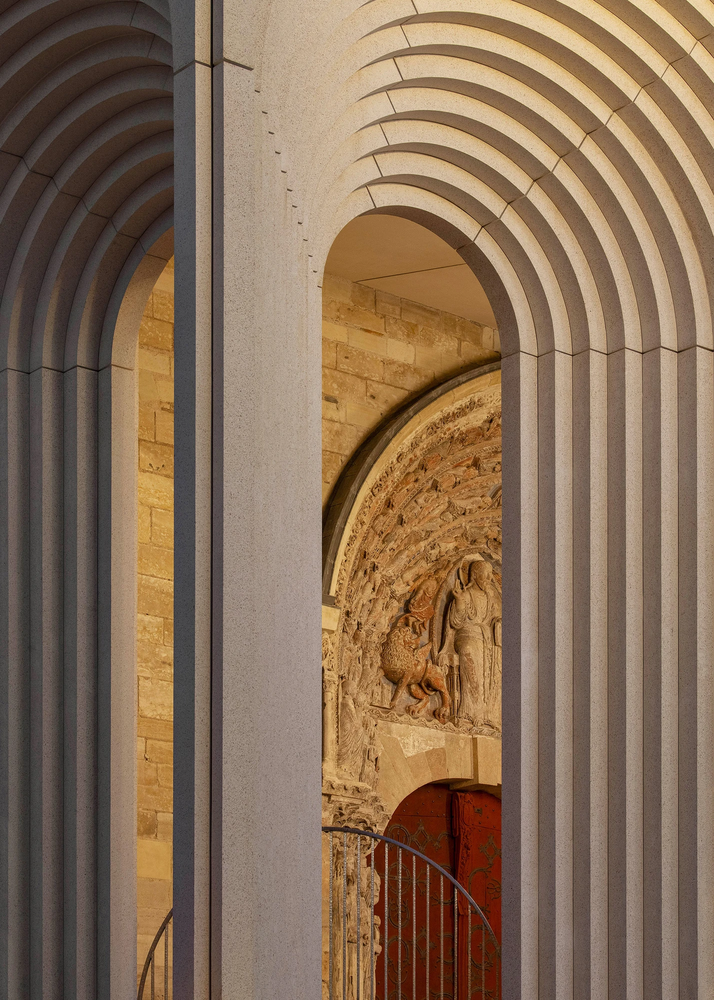 Angers Cathedral Entrance by Kengo Kuma and Associates — Oblique close-up at dusk: warm amber light from the enclosed nave spills through an archway, illuminating carved medieval stonework against pale cast concrete.