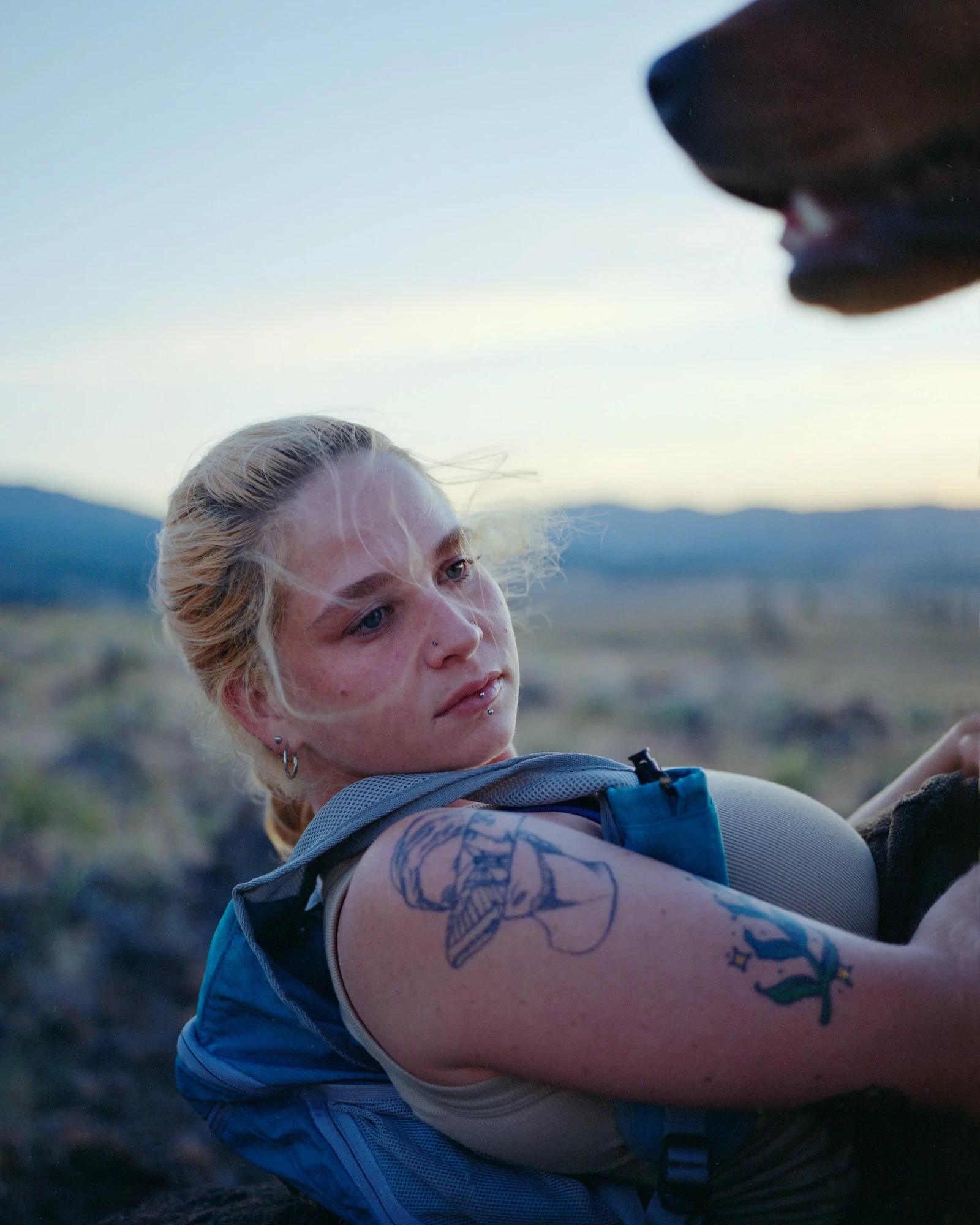 Camp by Blake Masi, photographed by Blake Masi — Portrait at dusk: a young woman with a tattoo on her arm looks down at a brown horse whose muzzle enters the frame from above right, mountains soft in the background.