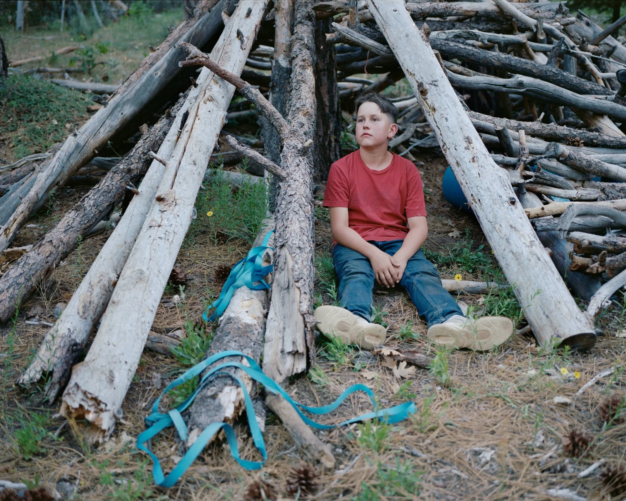 Camp by Blake Masi, photographed by Blake Masi — A boy in a red T-shirt sits against a pile of stacked grey logs, expression inward, a strip of blue plastic tarp on the pine-needle ground before him.