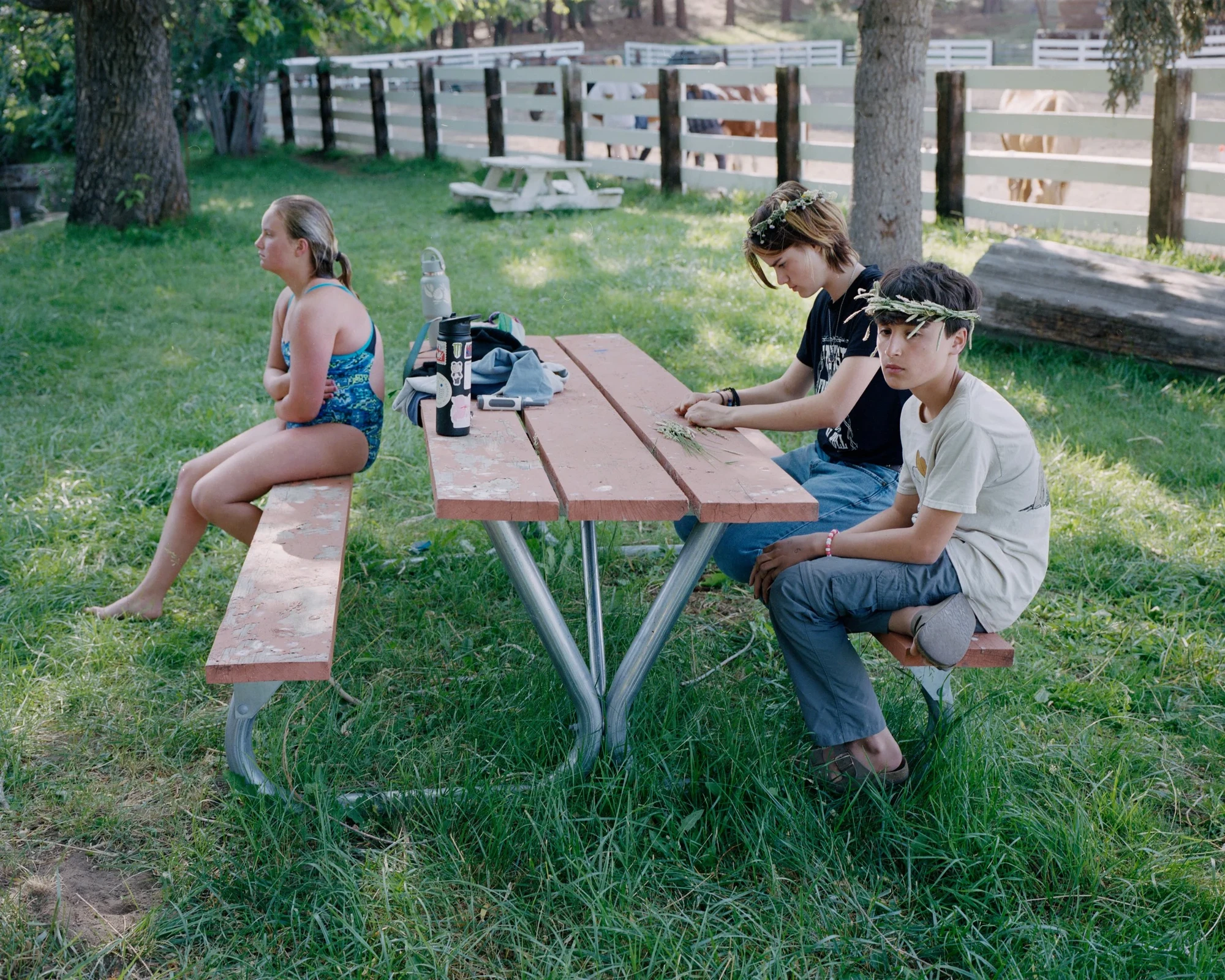 Camp by Blake Masi, photographed by Blake Masi — Three teenagers at a red-painted picnic bench on a grass lawn: one facing away in a swimsuit, two others side by side absorbed in their own objects.