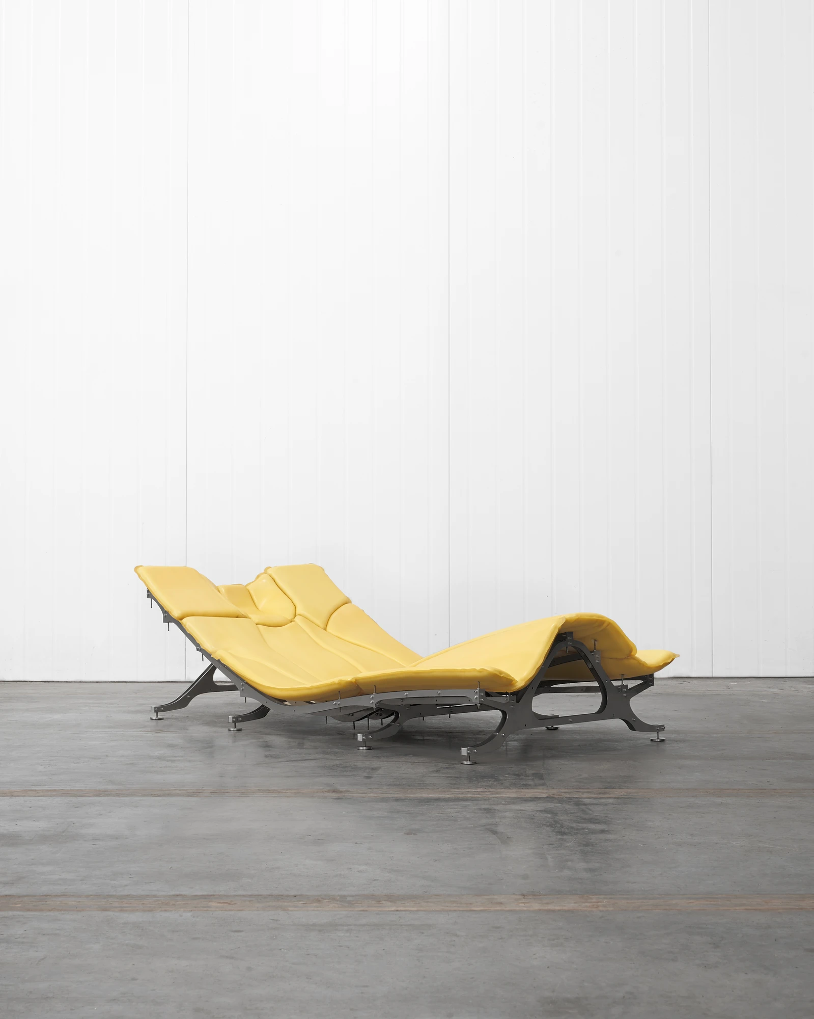 Daydreamer by onlyonly.studio, photographed by onlyonly studio — Daydreamer in three-quarter view on a concrete floor: a wide S-curved form in pale cadmium-yellow latex over a stainless steel eight-leg chassis, hovering centimetres above the ground.