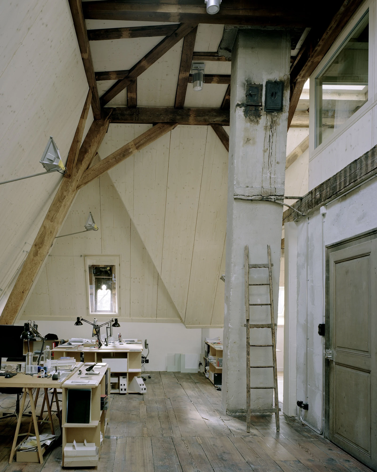 Ehemaliges Pfarrhaus Elisabethenstrasse by Vecsey Schmidt Architekten, photographed by Barbara Buhler — Large attic studio with the full king-post roof geometry exposed, whitewashed purlins, stone column, and timber ladder.