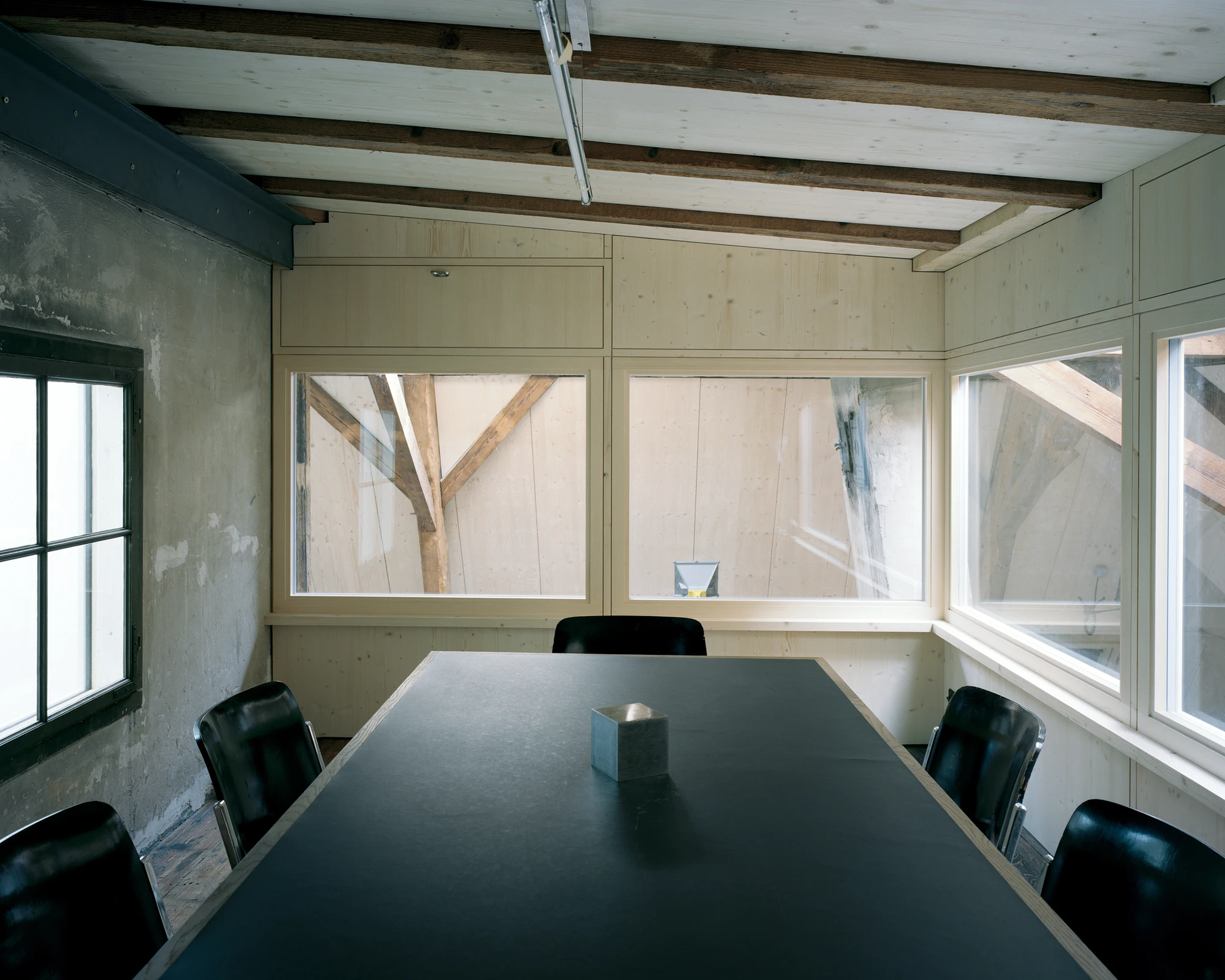 Ehemaliges Pfarrhaus Elisabethenstrasse by Vecsey Schmidt Architekten, photographed by Barbara Buhler — Attic meeting room with dark conference table, leather chairs, and plywood-framed skylights revealing oak trusses through translucent panels.