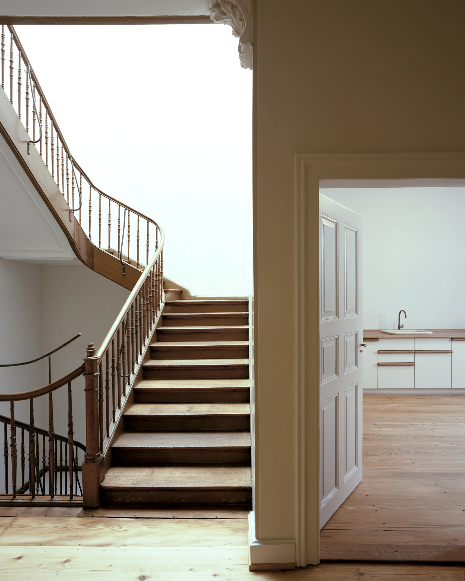 Ehemaliges Pfarrhaus Elisabethenstrasse by Vecsey Schmidt Architekten, photographed by Barbara Buhler — Original spiral staircase with dark wood balustrade; white kitchen with pine floor visible through the open door.