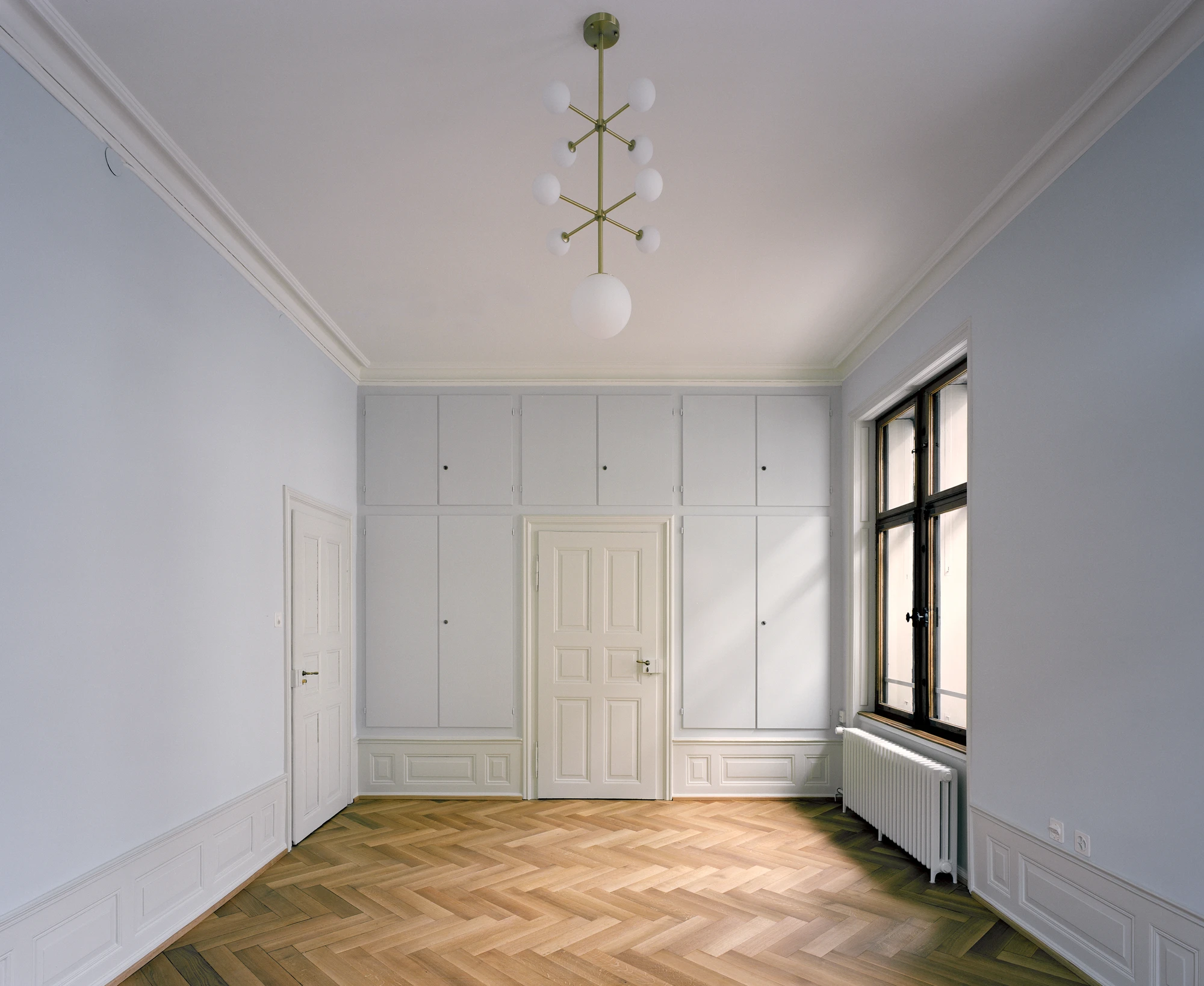 Ehemaliges Pfarrhaus Elisabethenstrasse by Vecsey Schmidt Architekten, photographed by Barbara Buhler — Empty bel etage room with herringbone parquet, panelled built-in cabinetry, brass sputnik pendant, and original moulded cornice.
