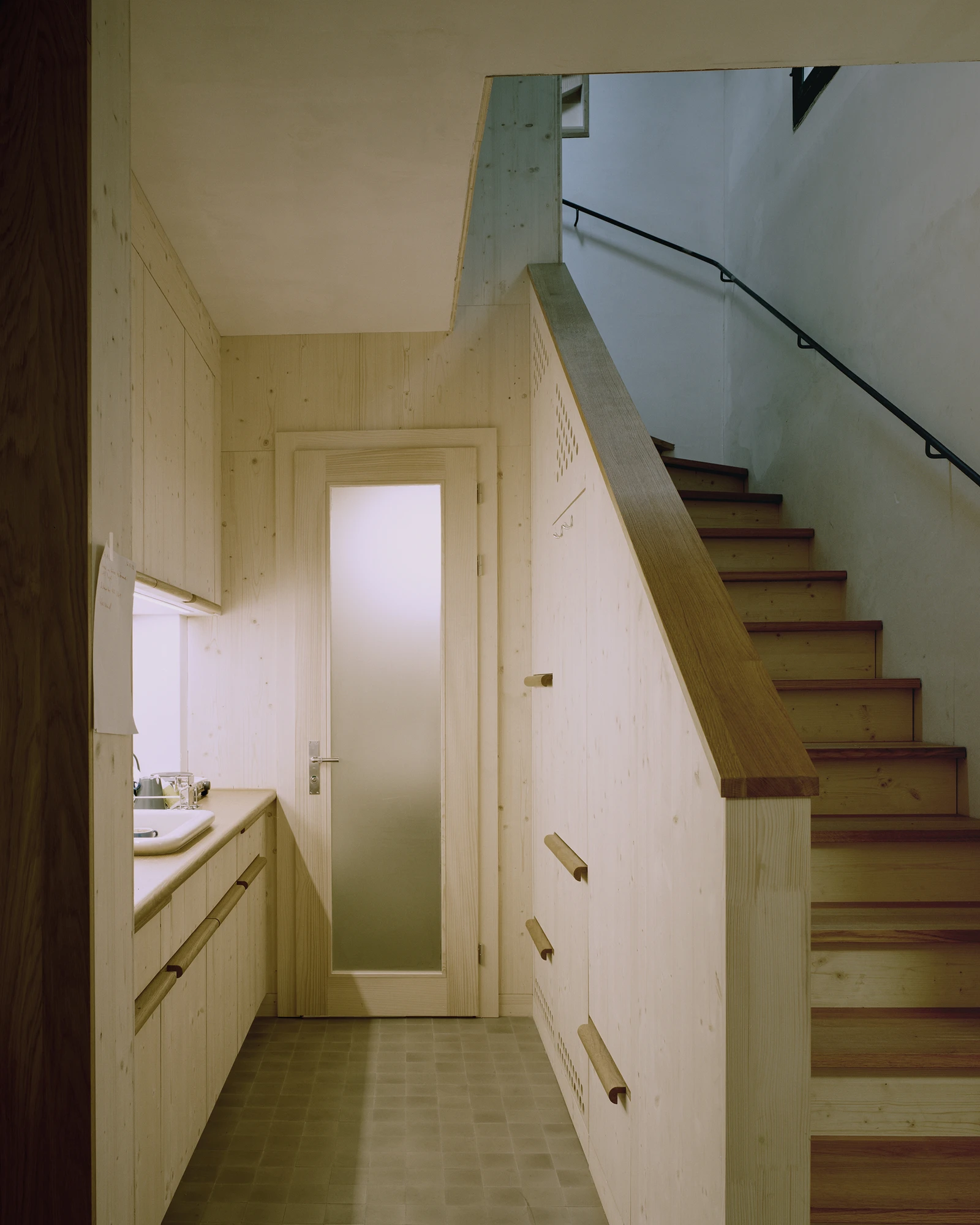 Ehemaliges Pfarrhaus Elisabethenstrasse by Vecsey Schmidt Architekten, photographed by Barbara Buhler — Attic corridor clad in raw spruce plywood with compact kitchen under the stair and frosted-glass door.