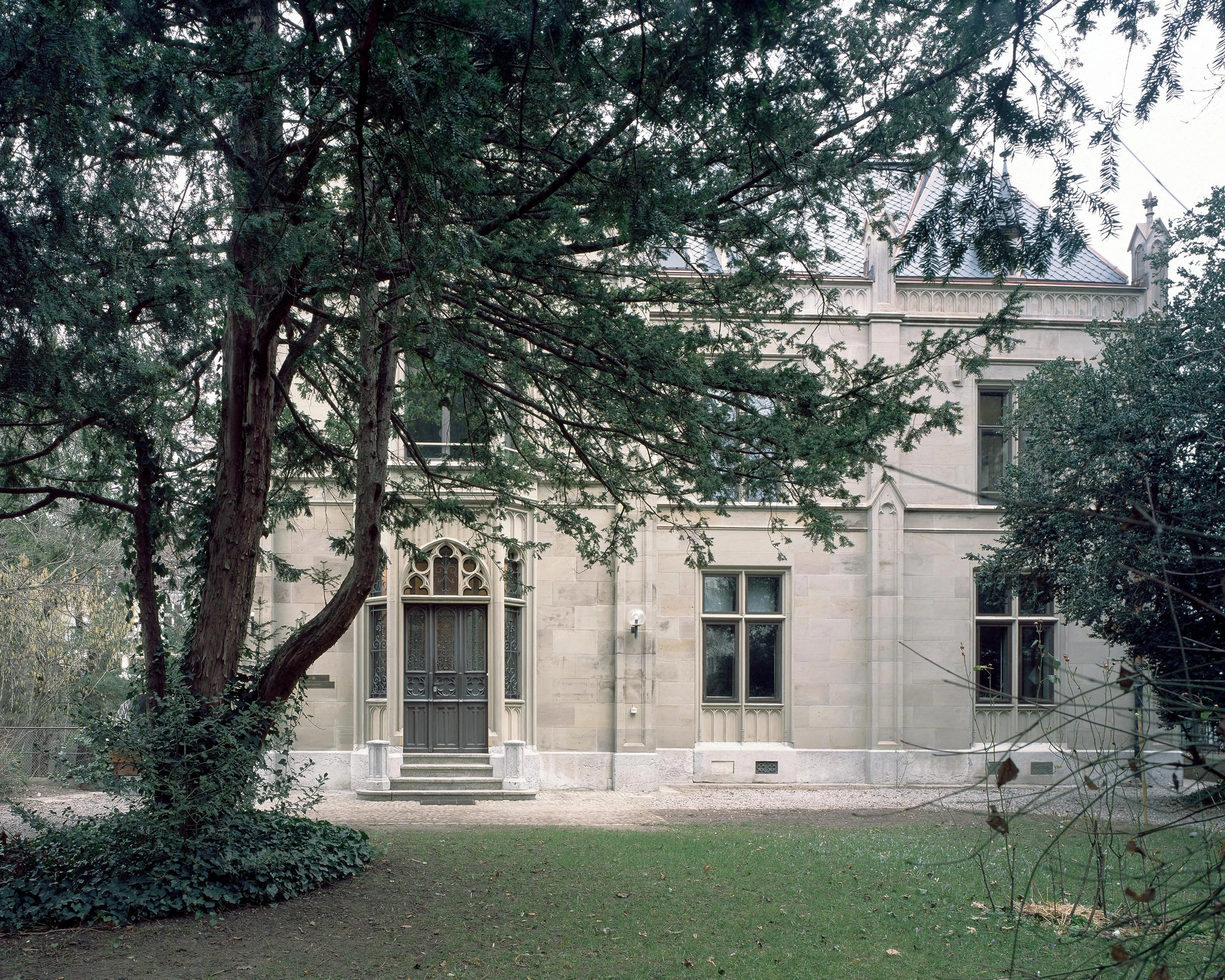 Ehemaliges Pfarrhaus Elisabethenstrasse by Vecsey Schmidt Architekten, photographed by Barbara Buhler — Garden view of the sandstone rectory with mature conifer and Gothic tracery entrance, parish garden foreground.