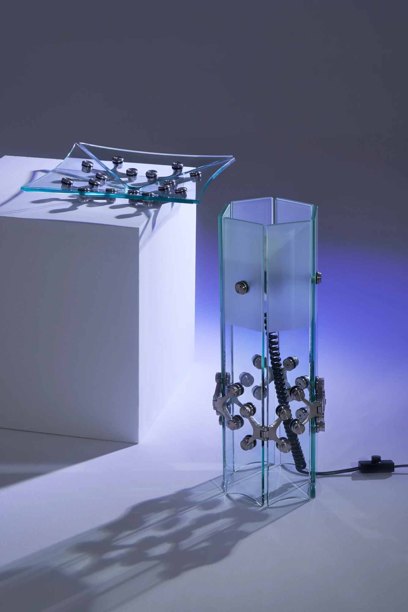 Ferric Glass by Tino Seubert, photographed by Piercarlo Quecchia, DSL Studio — Table lamp and flat glass bowl with spider hinges on a white plinth, blue ambient light, the panels showing the green-tinted high-iron glass edges.