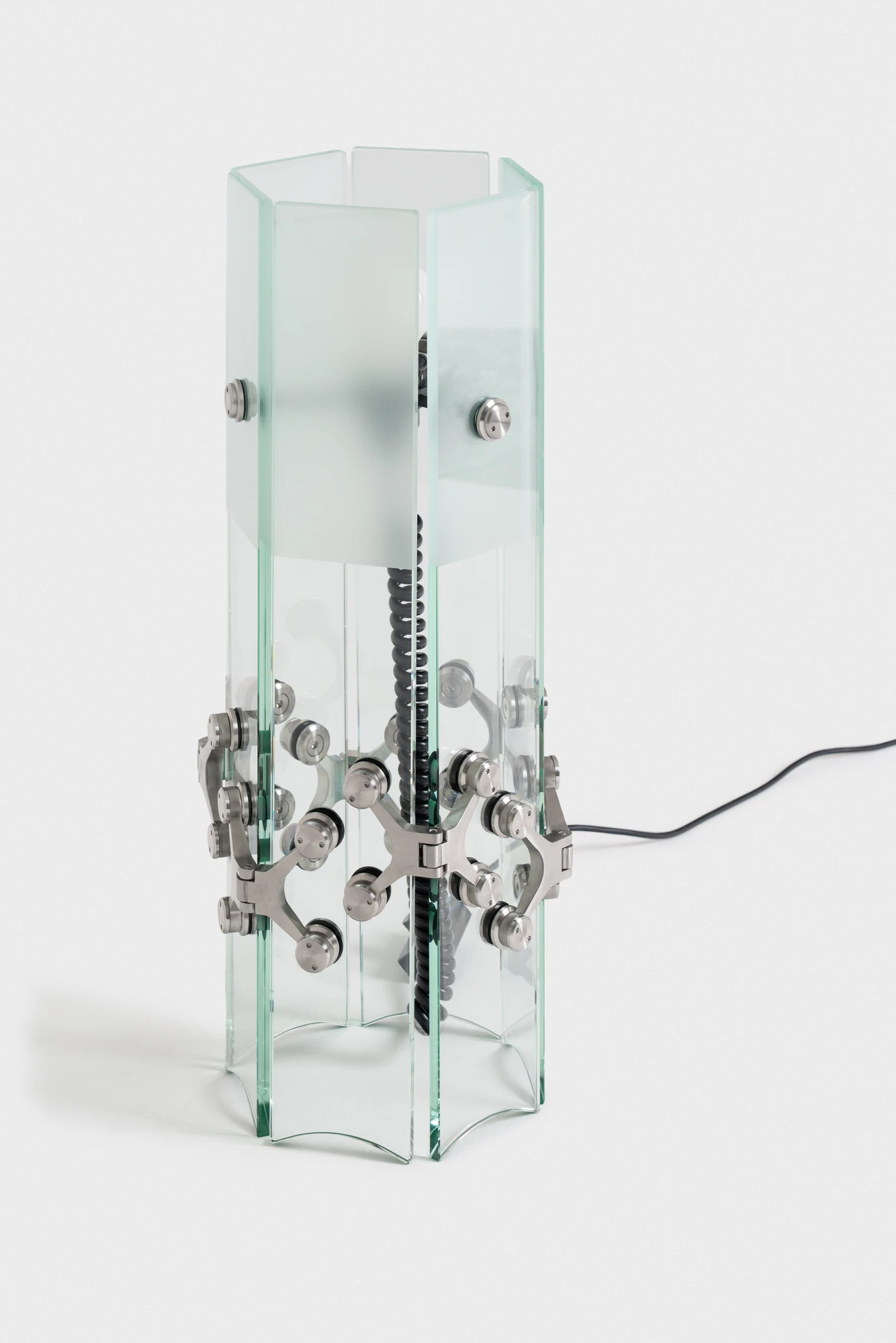 Ferric Glass by Tino Seubert, photographed by Piercarlo Quecchia, DSL Studio — Close detail of coil spring hardware between two glass panels, the stainless spring and spider hinge seen edge-on, glass edges showing the deep green iron tint.