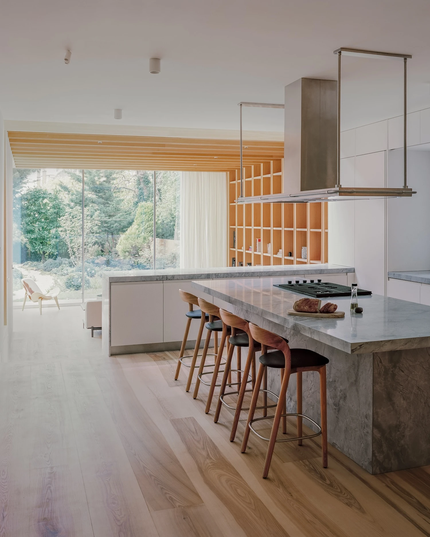 Panoramic House by MATA Architects, photographed by Nick Dearden — Kitchen island in grey-veined marble, walnut bar stools lined up beside it, the living room and garden glazing visible in the background through an open plan.