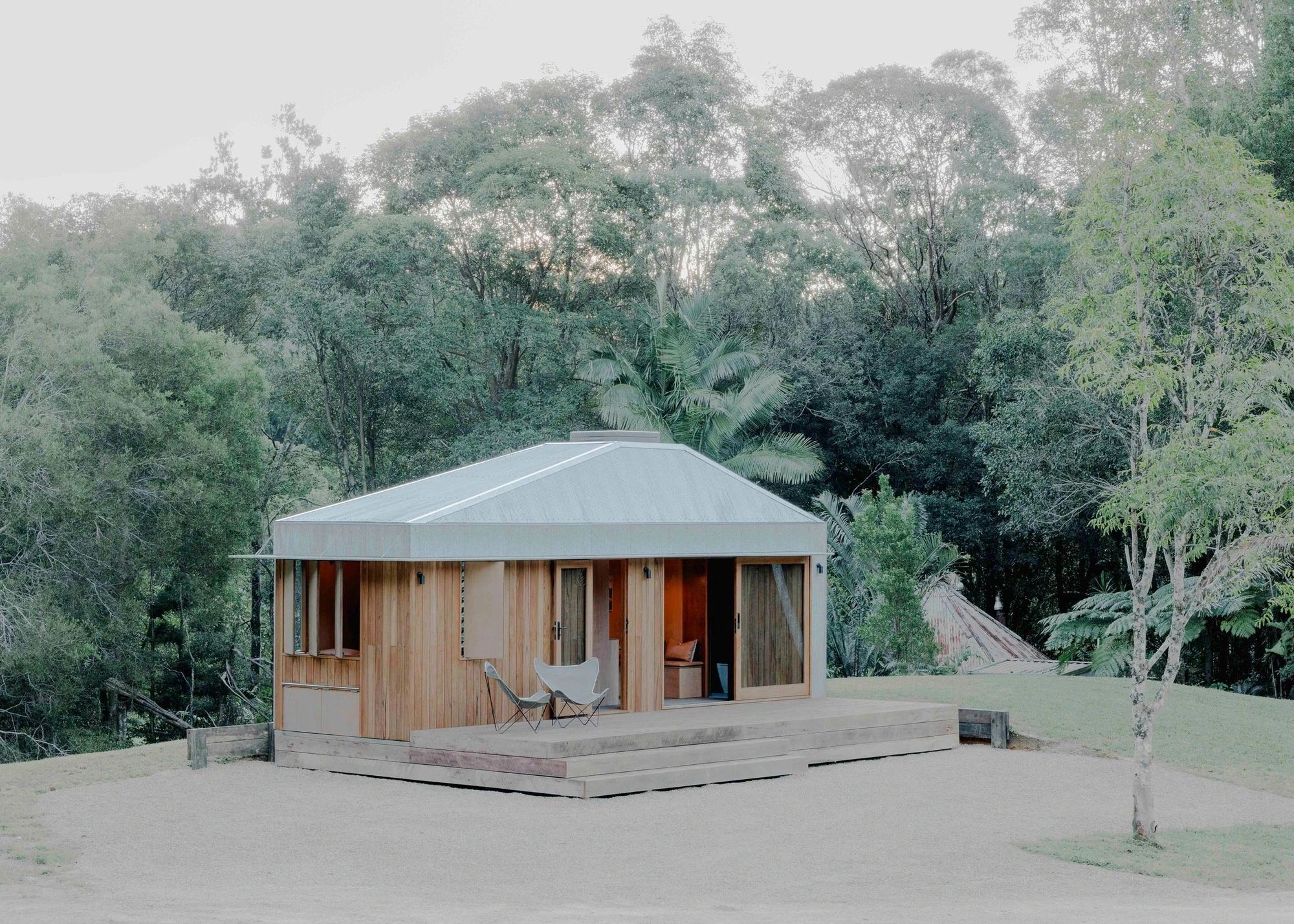 The Oculus by Hayley Pryor — The Oculus cabin from the front: a corrugated zinc hip roof with a central skylight apex, vertical timber-clad walls, two chairs on a low timber deck facing eucalyptus forest.