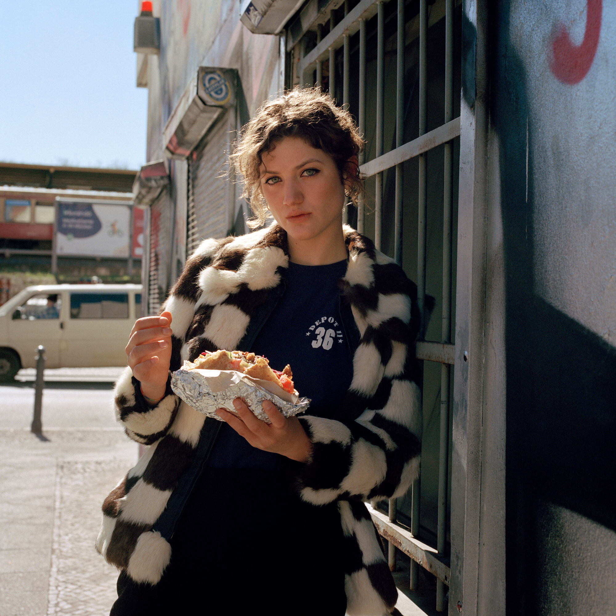 Capital Daughters by Lucia Jost, photographed by Lucia Jost — Woman in a black-and-white check faux-fur coat holding a street-food wrap on a Berlin pavement, direct eye contact.