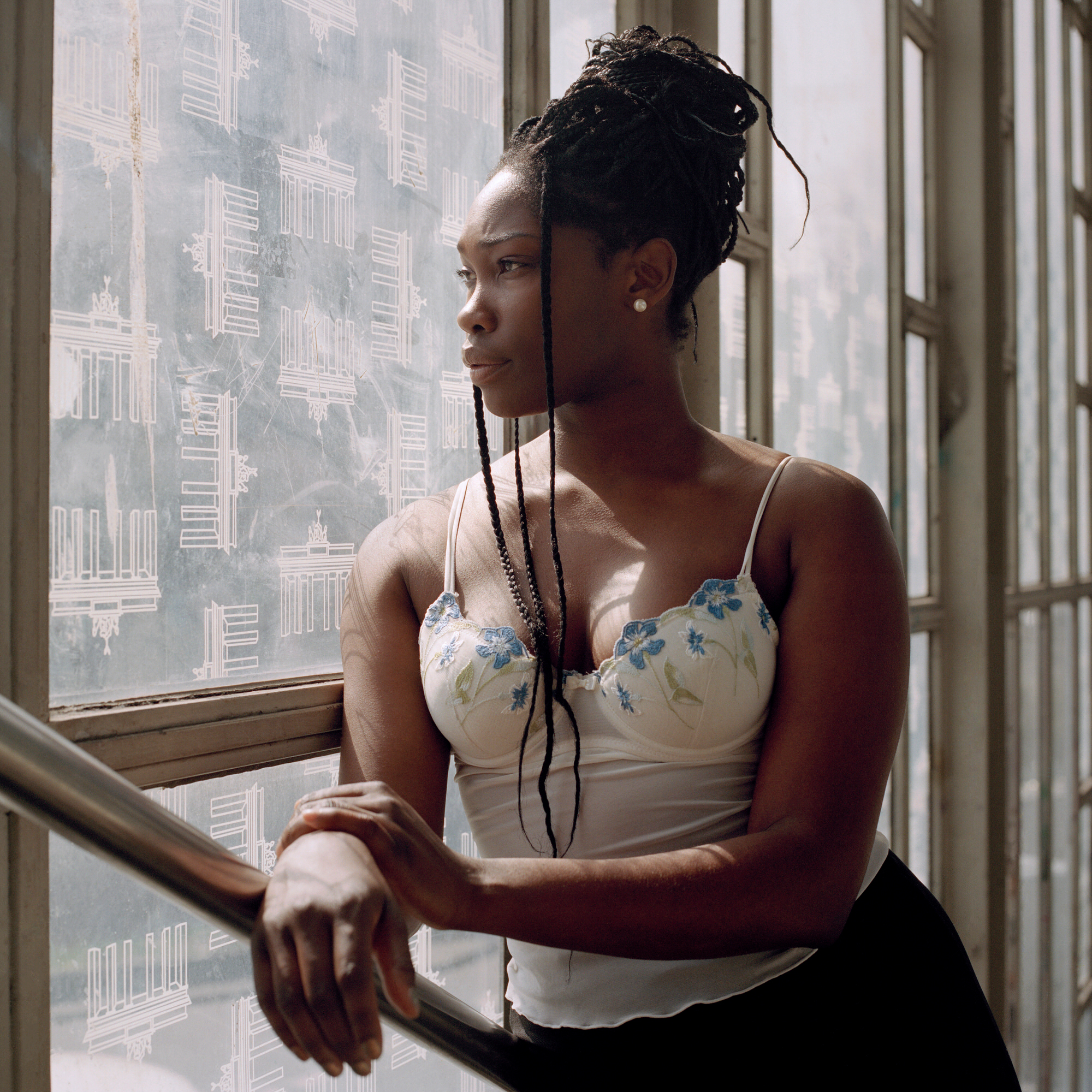 Capital Daughters by Lucia Jost, photographed by Lucia Jost — Woman in a floral corset top gazes out through a large window, Berlin buildings visible below, soft natural light.