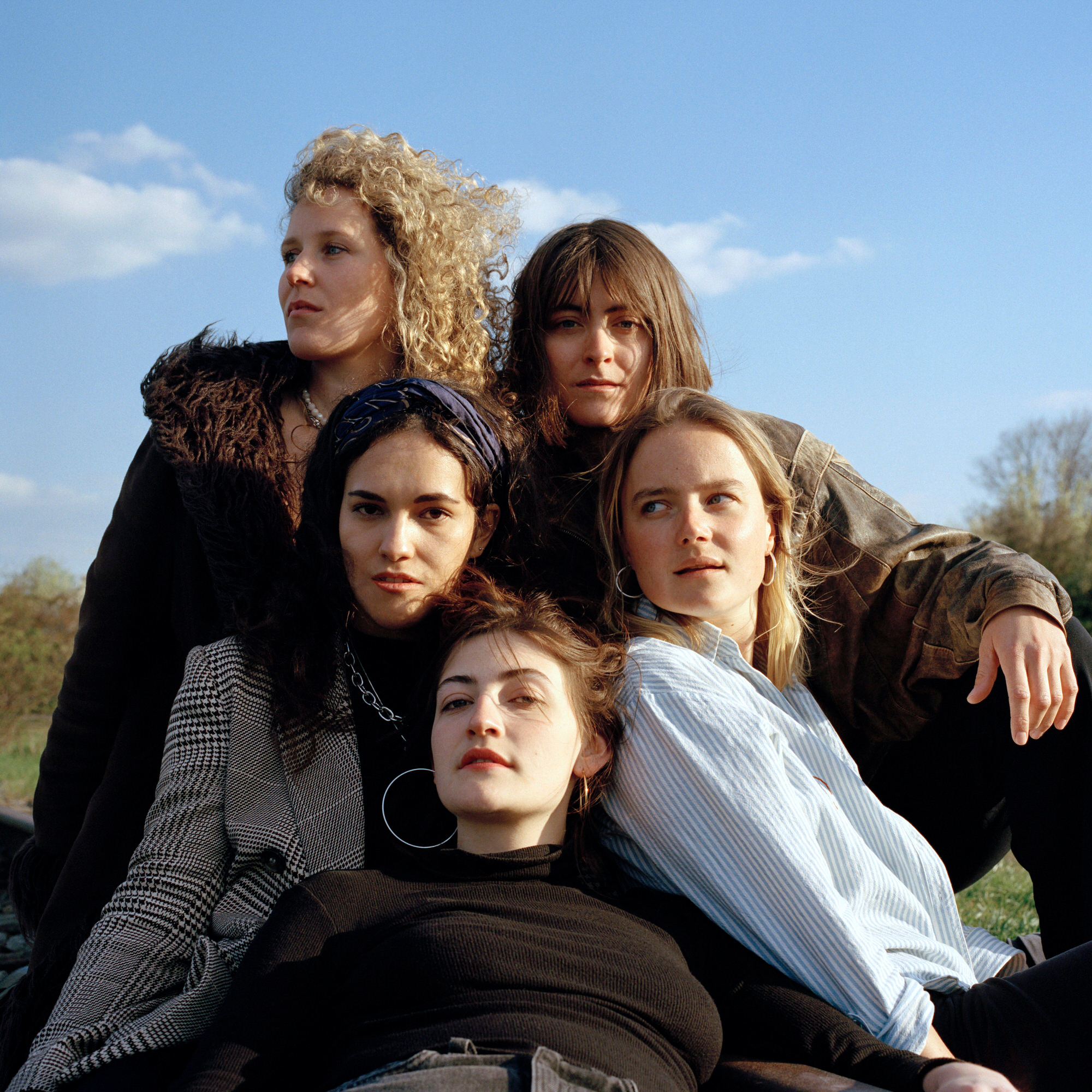Capital Daughters by Lucia Jost, photographed by Lucia Jost — Five women outdoors in an open field, grouped in a loose pyramid, a range of coats and jackets, clouds behind.
