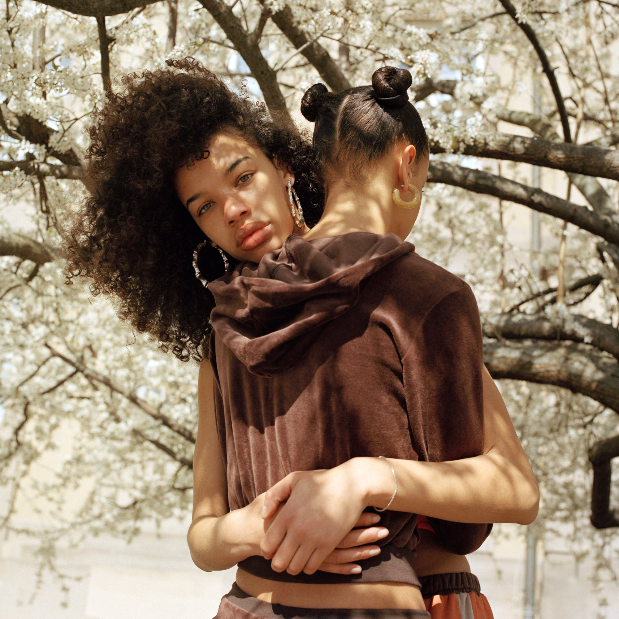 Capital Daughters by Lucia Jost, photographed by Lucia Jost — Two women embrace under blooming trees, one looking at the camera, textured brown knitwear, spring light.