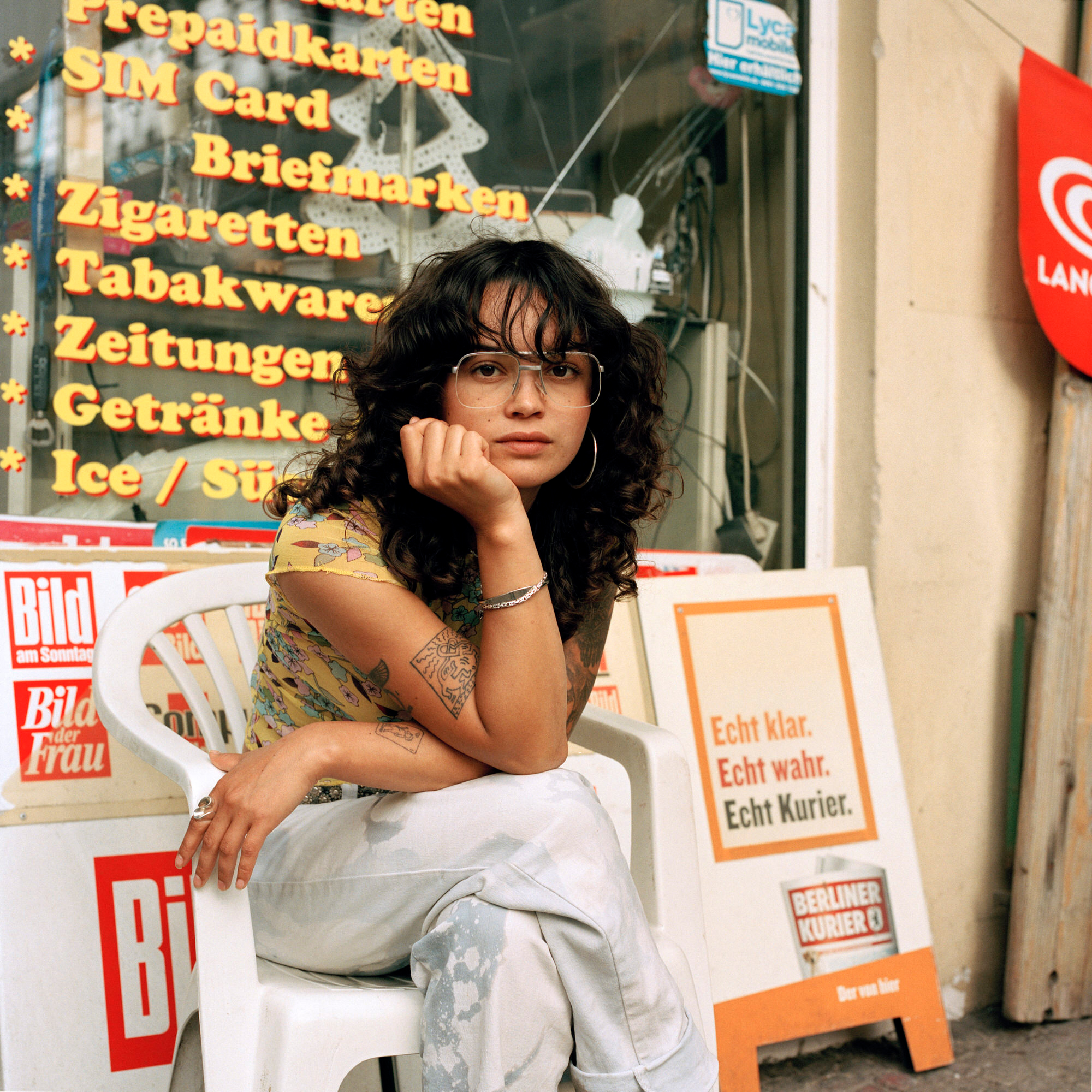 Capital Daughters by Lucia Jost, photographed by Lucia Jost — Woman sitting outside a newsagent with Bild and Berliner Kurier displayed, curly hair, layered jewellery, resting chin on hand.