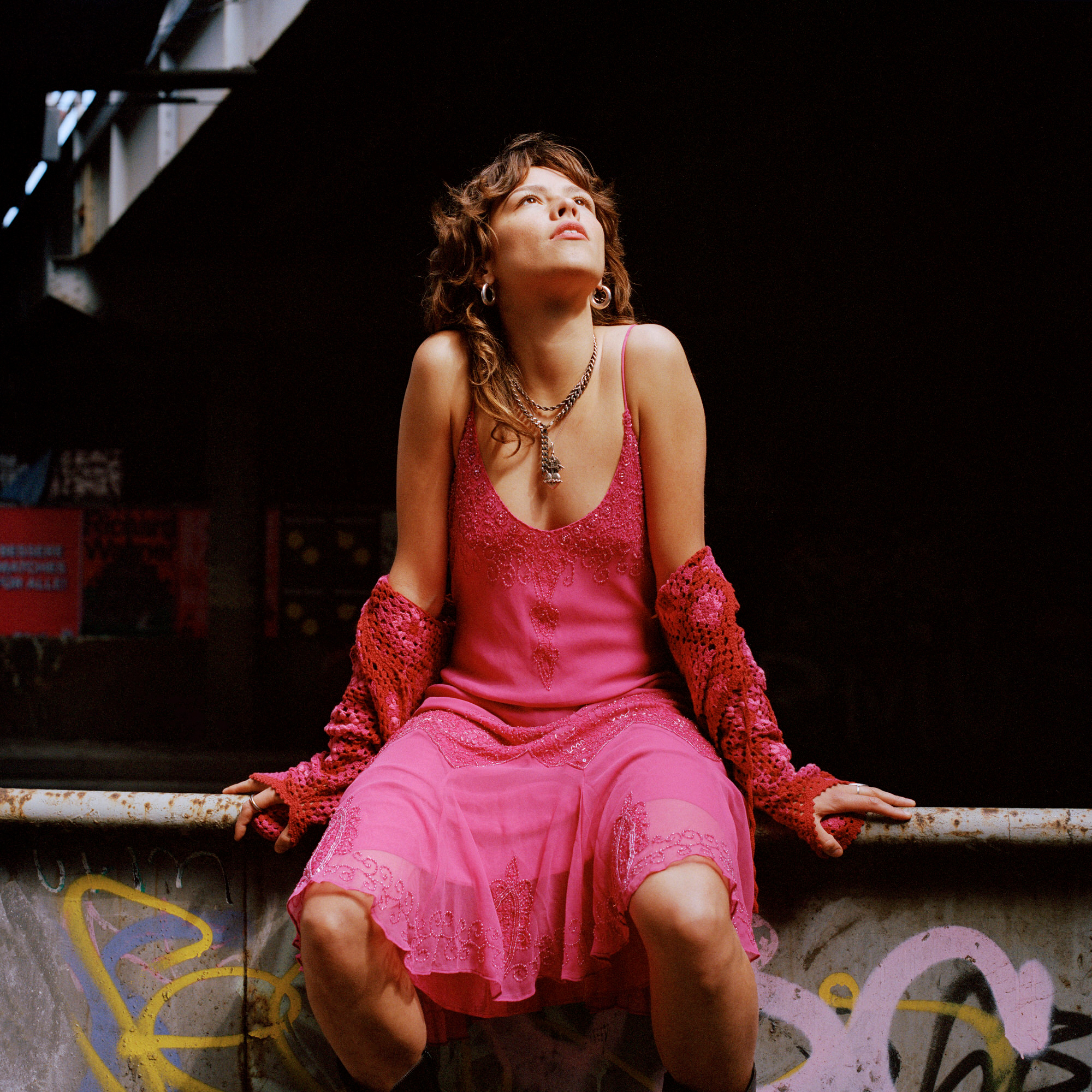 Capital Daughters by Lucia Jost, photographed by Lucia Jost — Woman in a pink lace slip dress seated on a graffitied metal ledge in a dark urban passage, looking upward.