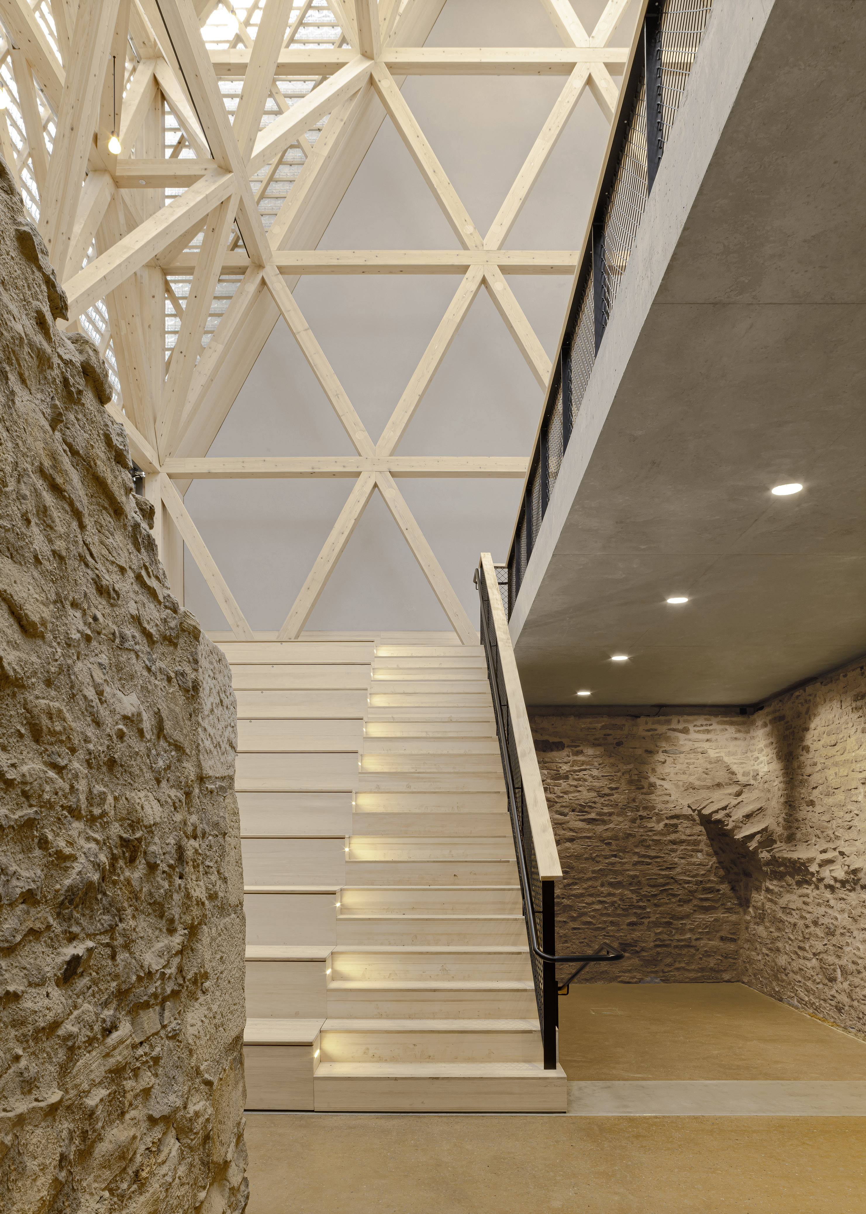 Historical Oberamteistraße Museum by wulf architekten, photographed by Brigida González — Interior staircase with lit treads descending past exposed rubble stone walls, the glass-and-timber roof structure visible above.