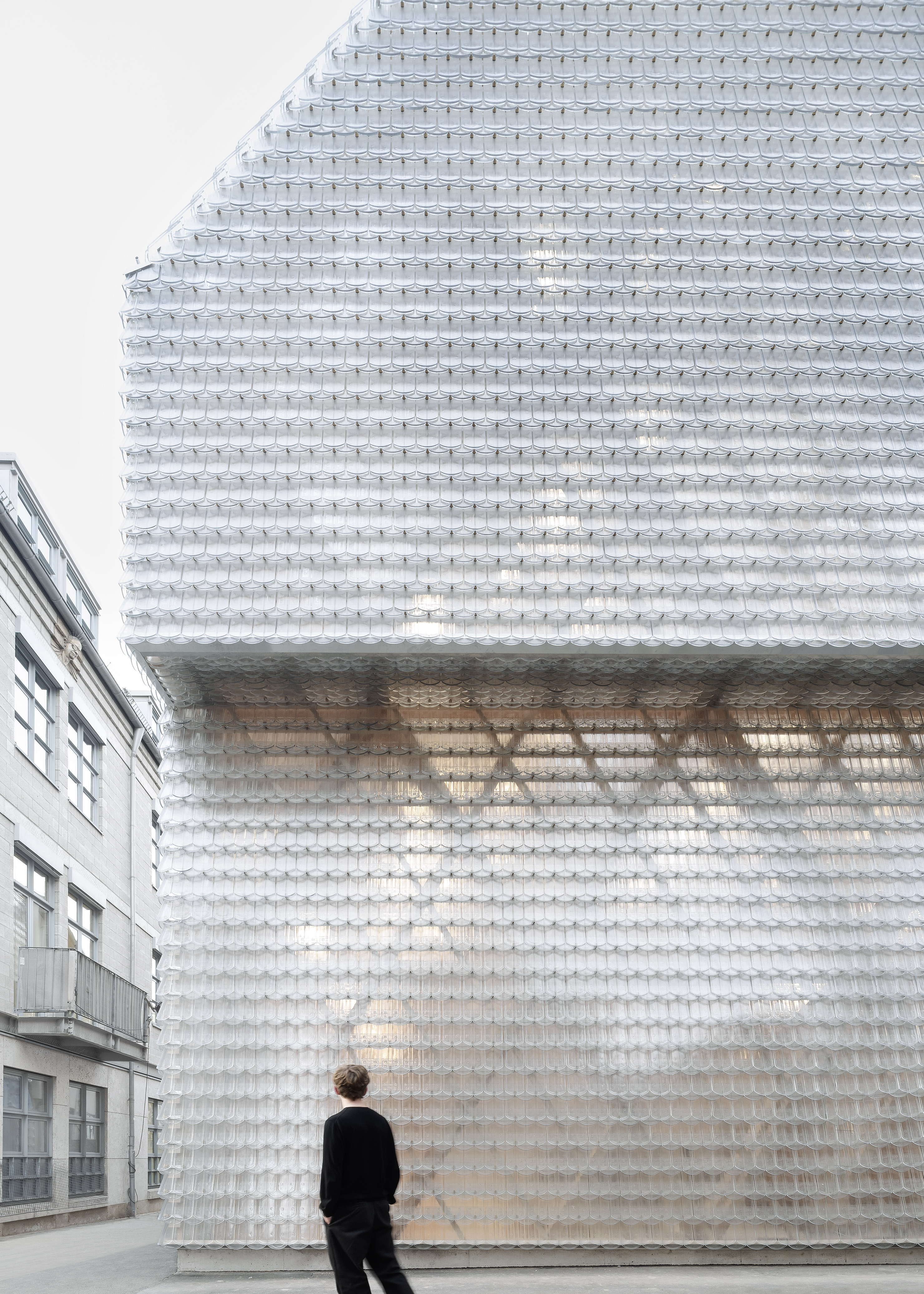 Historical Oberamteistraße Museum by wulf architekten, photographed by Brigida González — Close street elevation: the glass beaver-tail tile facade at full height, a figure in black standing at its base, the wooden truss faintly legible behind.