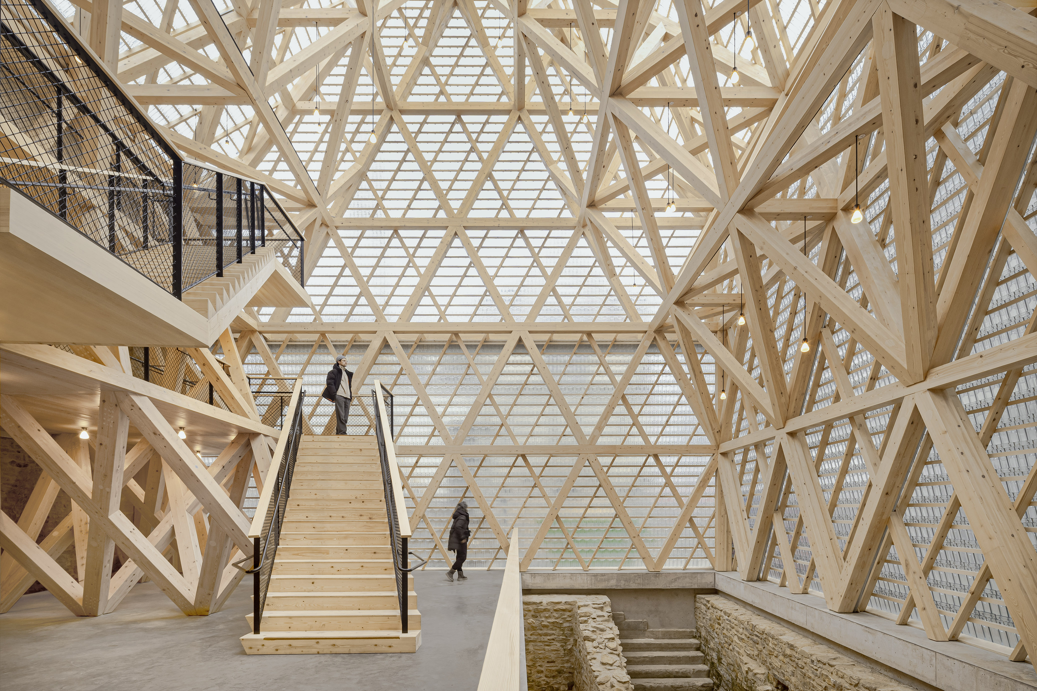 Historical Oberamteistraße Museum by wulf architekten, photographed by Brigida González — Interior main hall: pale spruce truss members form a latticework overhead as two visitors use the staircase above the medieval cellar.