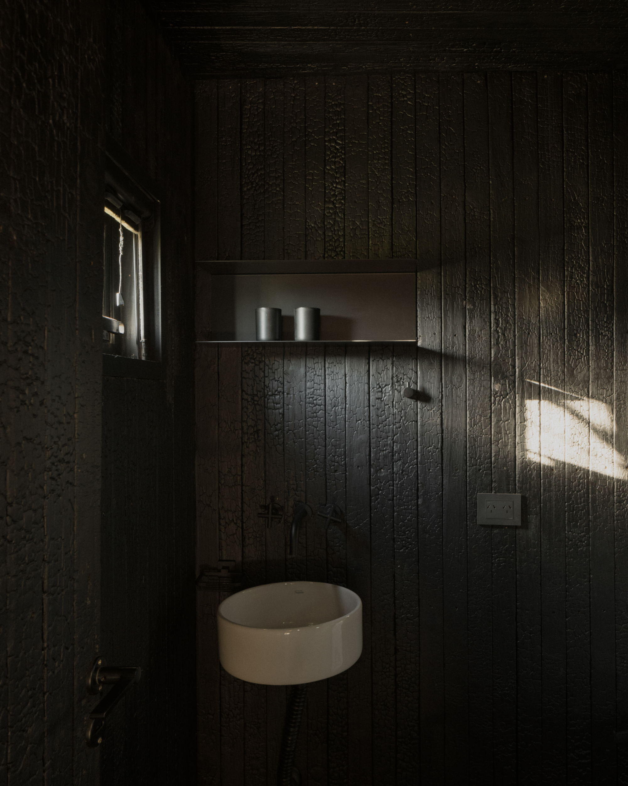Pavilion El Durazno by Nicolás Oks, photographed by Federico Cairoli — Bathroom in fully charred-timber cladding, single small window casting a bar of light across the sink and wall shelf.