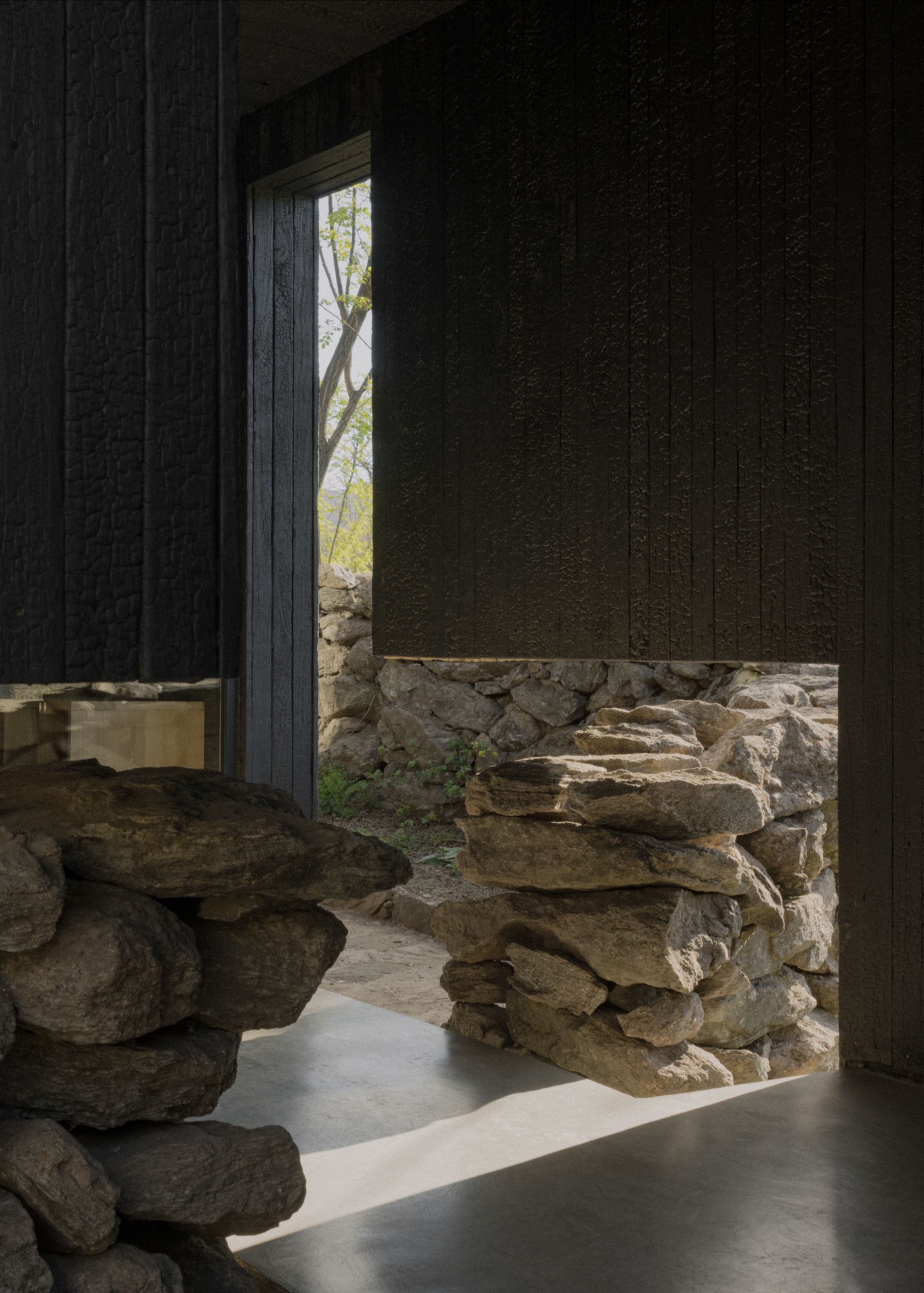 Pavilion El Durazno by Nicolás Oks, photographed by Federico Cairoli — Interior threshold view through a tall opening: the stone wall is framed in close-up, charred timber on either side, pale concrete floor.