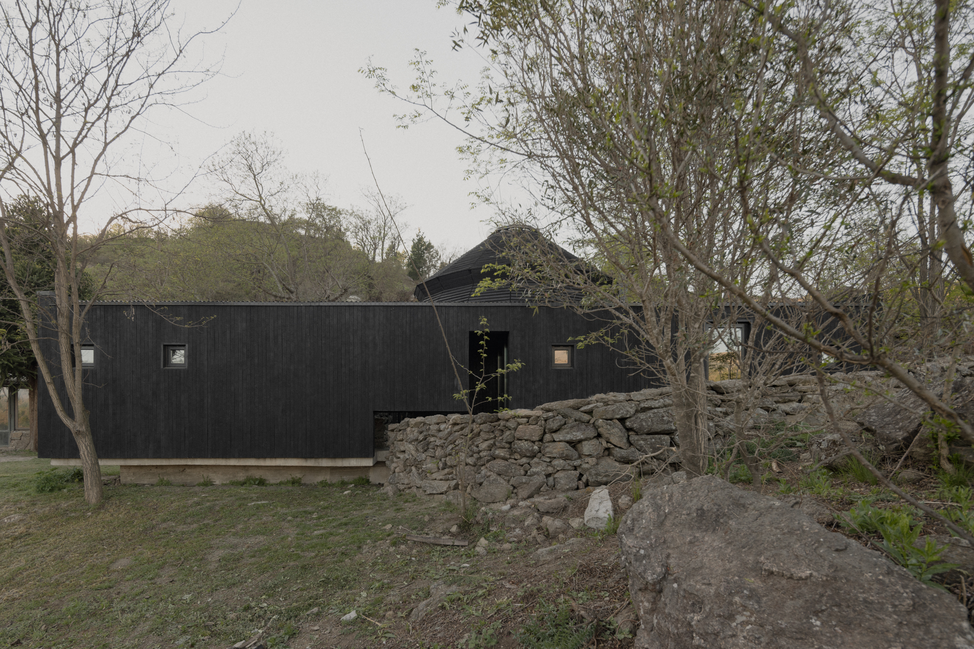 Pavilion El Durazno by Nicolás Oks, photographed by Federico Cairoli — The long black service prism with small square windows punched into charred-timber cladding, stone wall and bare trees framing the scene.