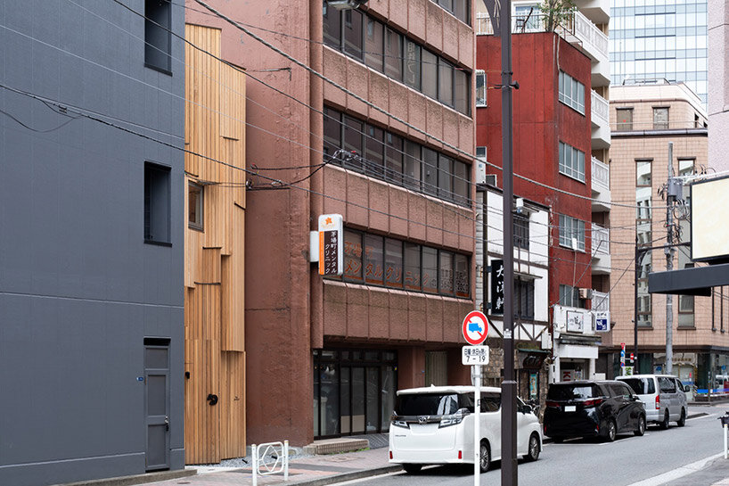 Prewood by VUILD, photographed by Tomoyuki Kusunose, Hayato Kurobe — Wide street shot showing the cedar building as a sliver between taller structures on either side, a pedestrian passing at the corner.