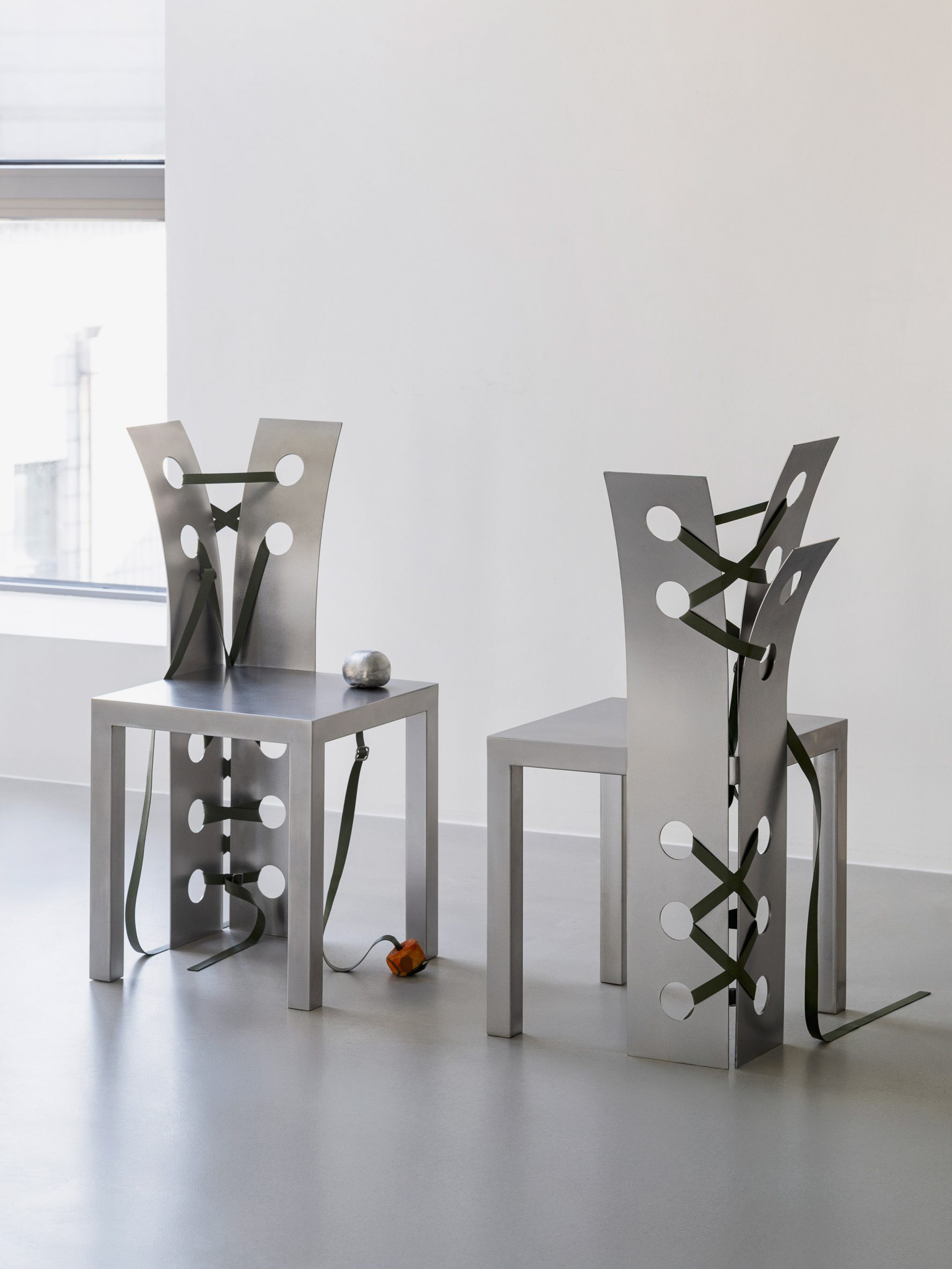 Pul Collection by Nara Lee, photographed by Namuk Kim — Two chairs with green leather straps facing a window, natural light catching the brushed steel surface and loose strap ends at their bases.
