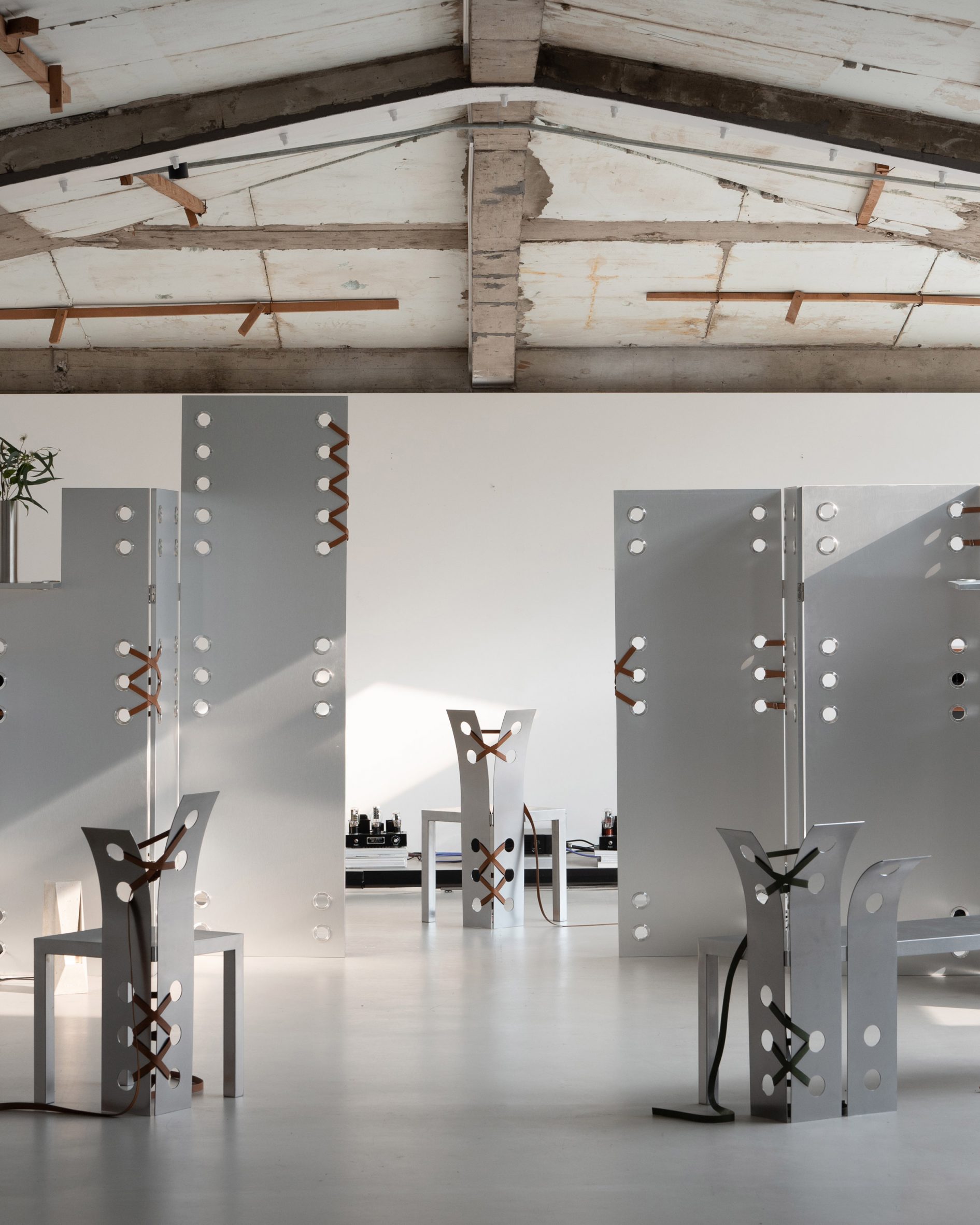 Pul Collection by Nara Lee, photographed by Namuk Kim — Exhibition view: chairs and tall folding screens with punched holes and leather lacing stand across a wide white-floored space beneath exposed timber beams.