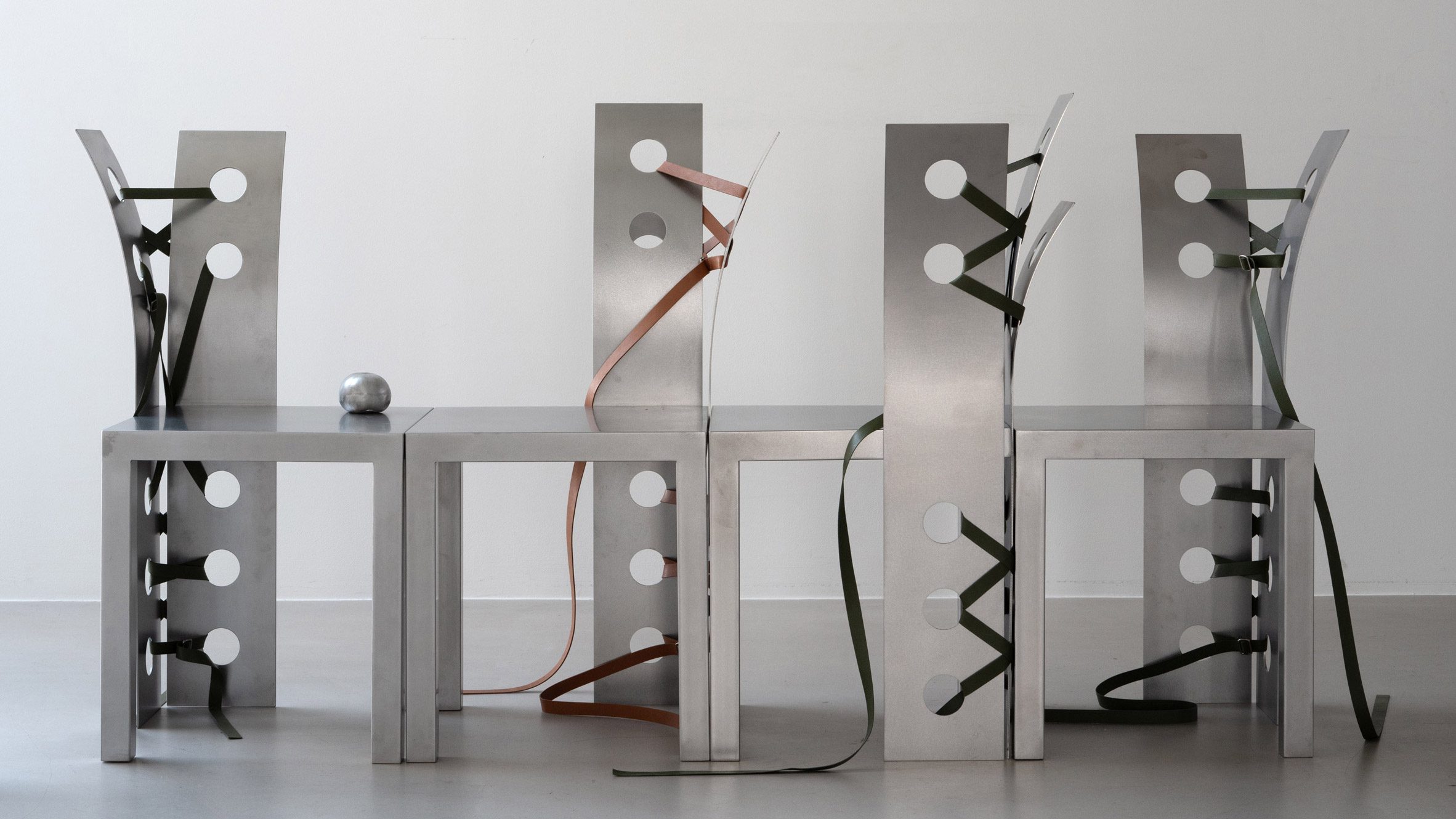 Pul Collection by Nara Lee, photographed by Namuk Kim — Four chairs in two facing pairs, tan and green leather straps threaded through their punched steel backs, a small silver sphere on one seat.