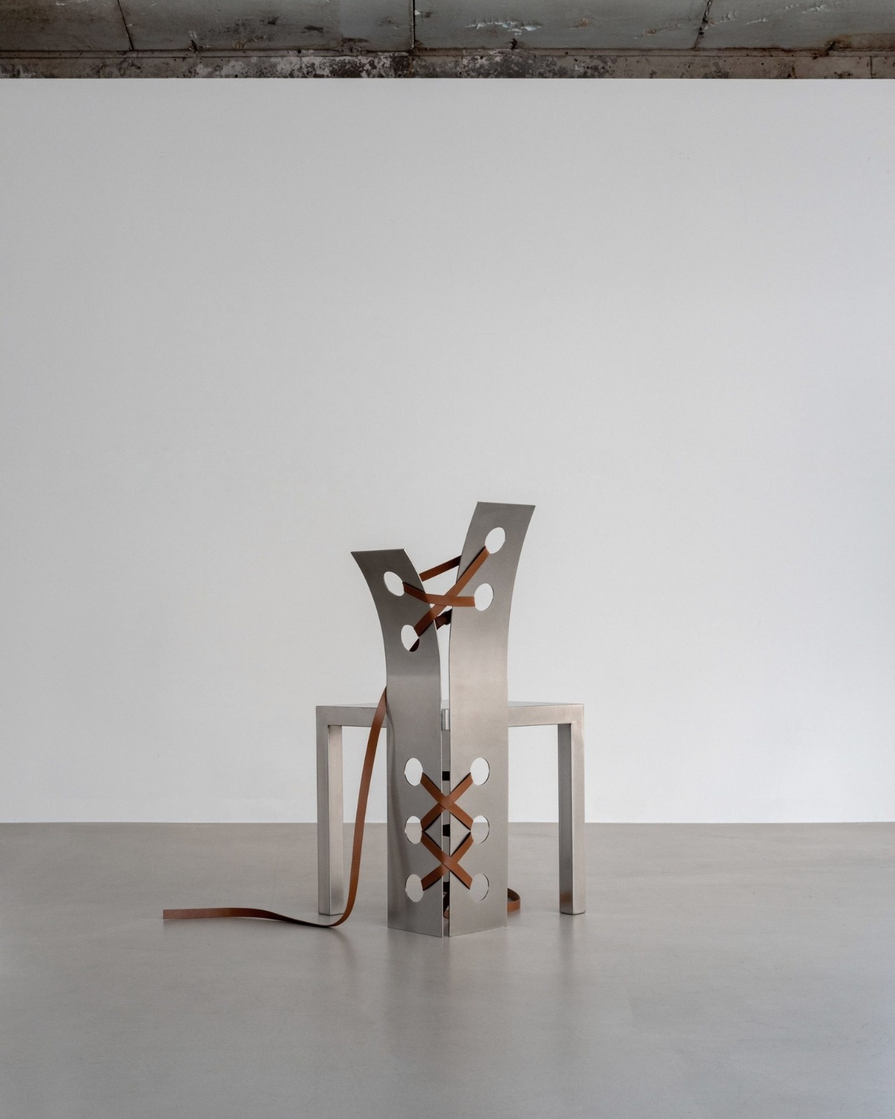 Pul Collection by Nara Lee, photographed by Namuk Kim — A single chair with tan leather lacing isolated in a white room beneath a raw concrete ceiling, strap trailing on the grey polished floor.