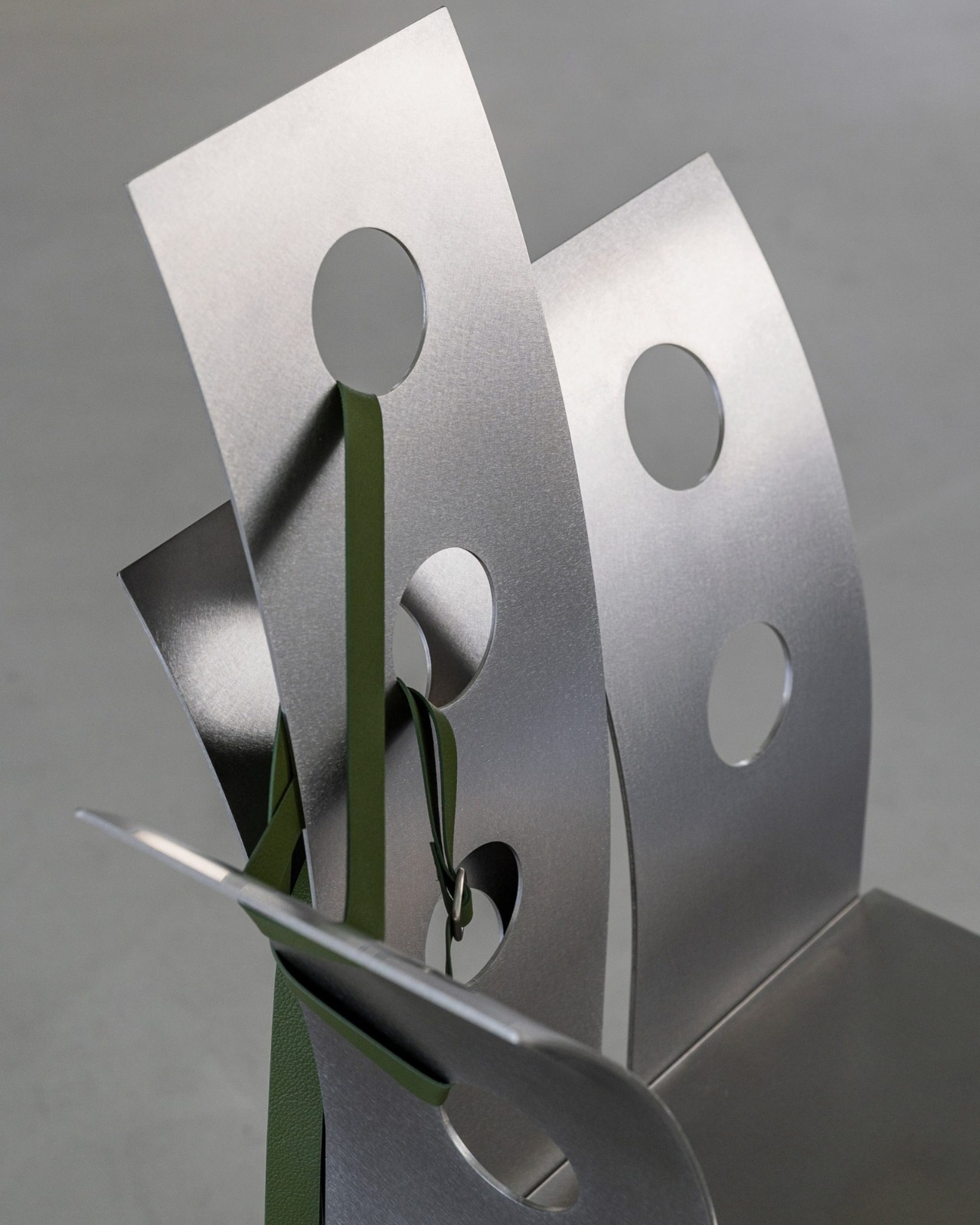 Pul Collection by Nara Lee, photographed by Namuk Kim — Close detail of the punched steel backrest: circular holes, green leather strap threaded in an X pattern, the strap buckle visible at the lower edge.
