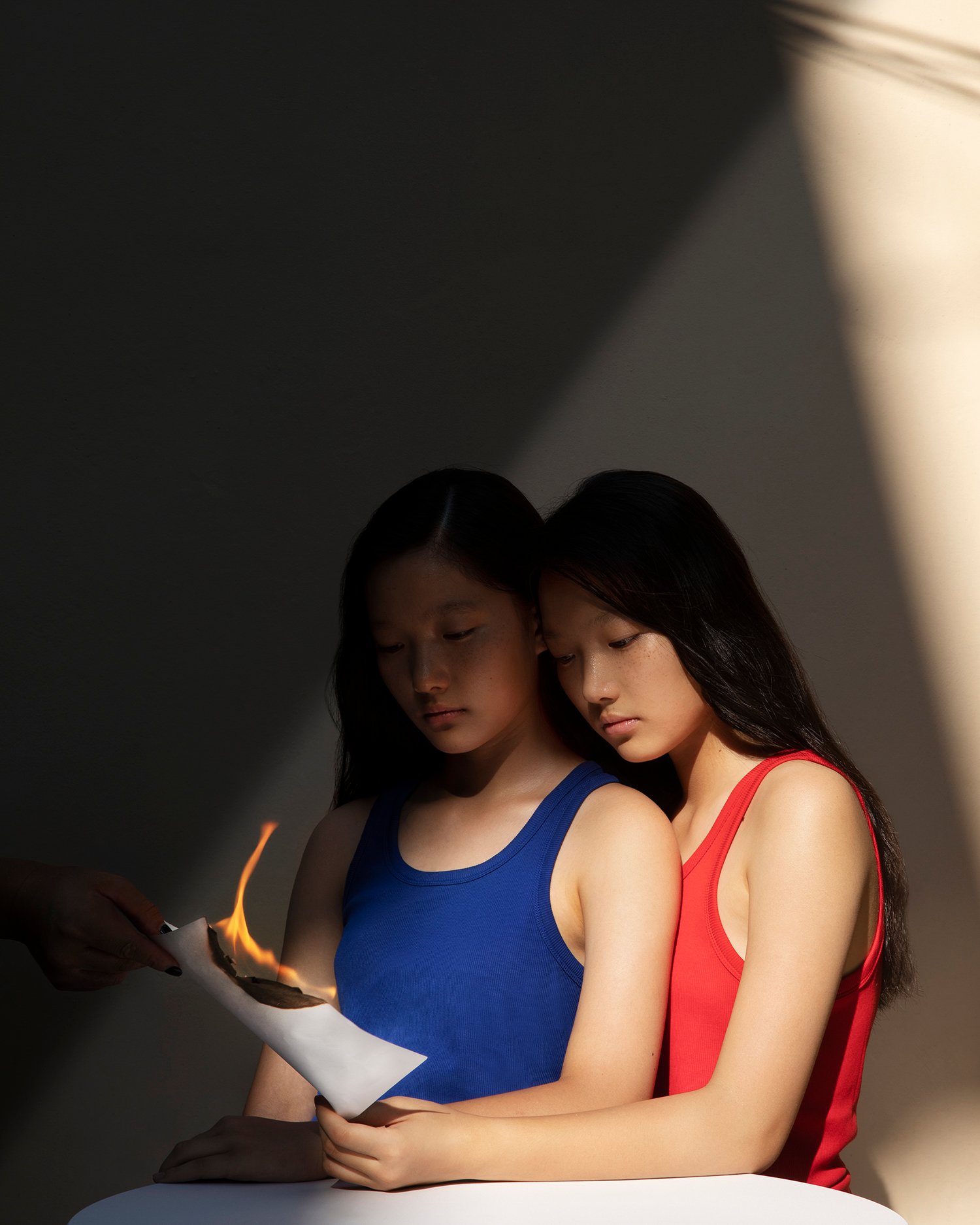 Trust Me by Jinyong Lian, photographed by Jinyong Lian — Two young women in blue and red tank tops lean together while a hand holds a burning page between them, diagonal light from above.