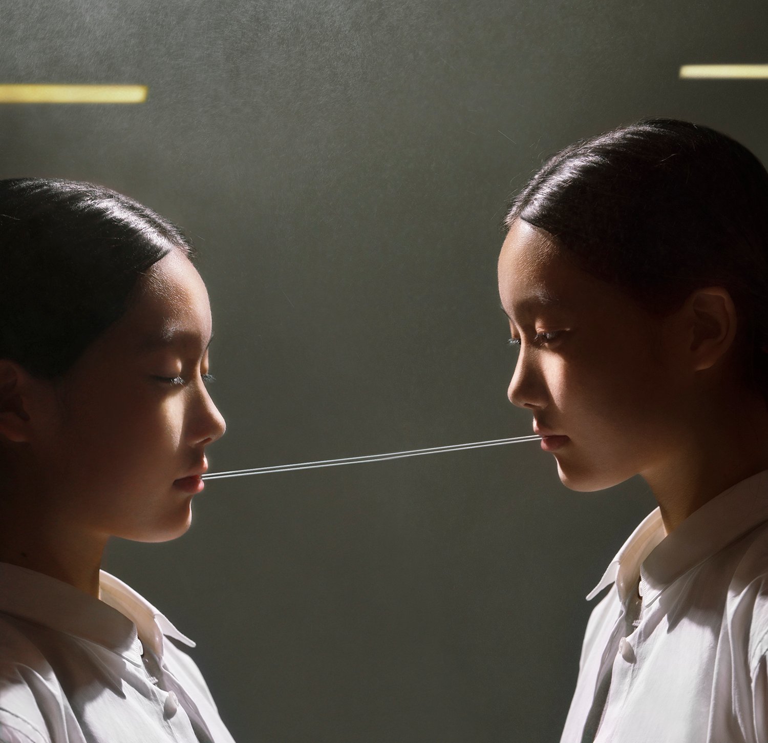 Trust Me by Jinyong Lian, photographed by Jinyong Lian — Two women in white shirts face each other with a single thin thread stretching from mouth to mouth between them.