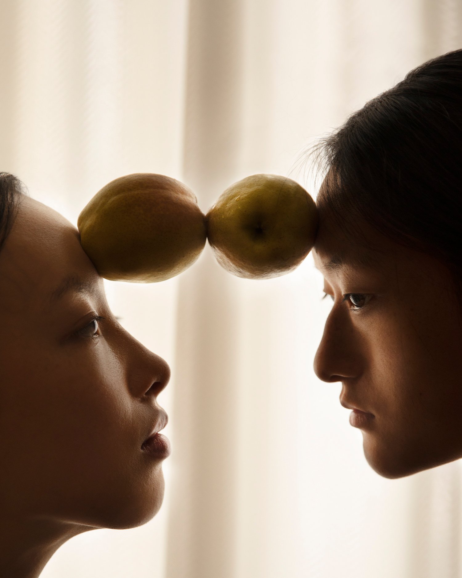Trust Me by Jinyong Lian, photographed by Jinyong Lian — Two profiles face each other in warm-toned shadow, a pear balanced between their foreheads at the temples.